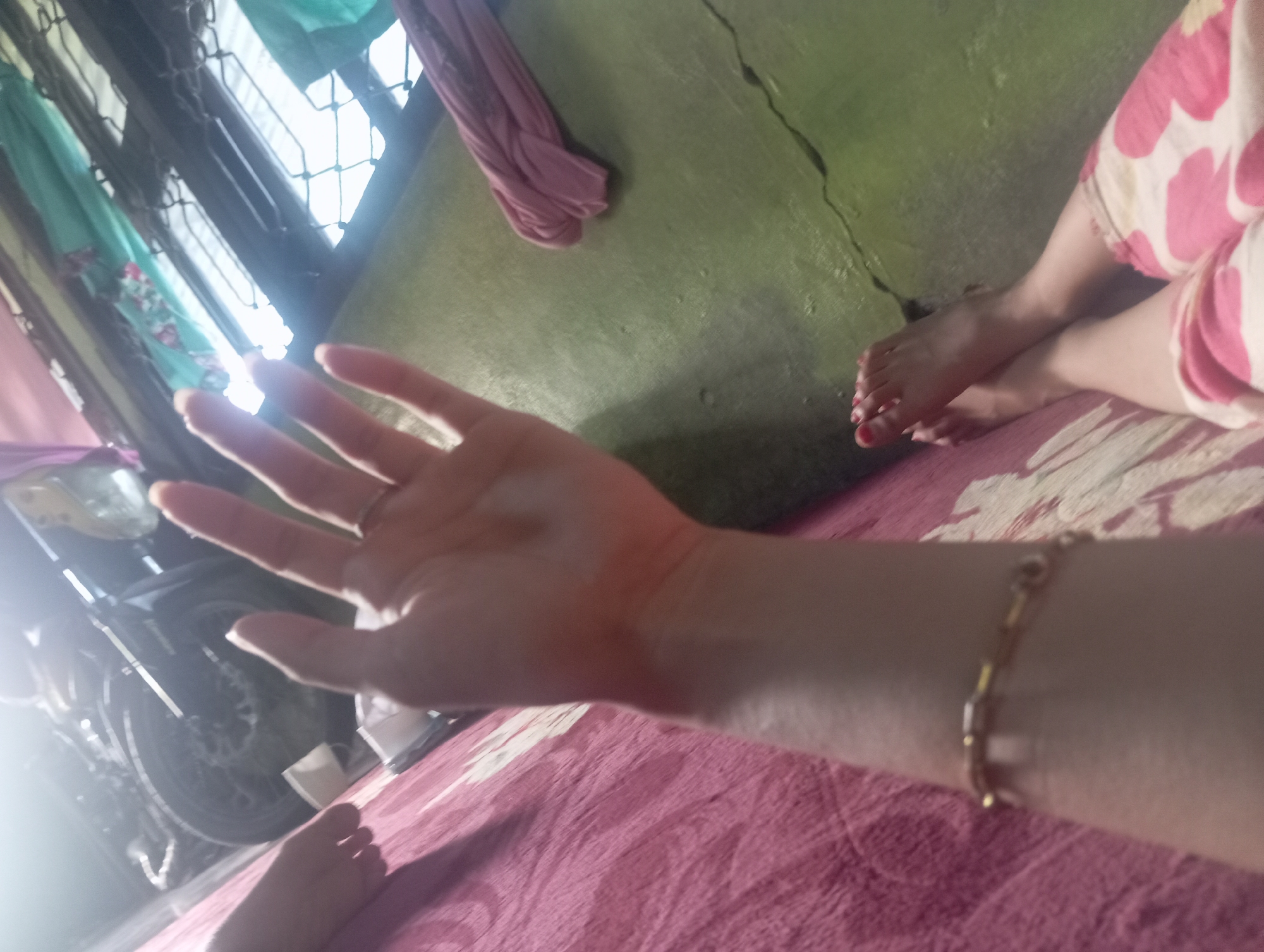 A person's hand is held up in the foreground, with their palm facing the camera. The hand appears to have some orange pigment on it, possibly henna or makeup. In the background, a motorcycle is partially visible, as are a pink cloth draped over a surface, and a wall painted green. Two feet, one with red nail polish, are visible in the lower right corner of the frame, resting on a pink patterned surface. The scene is indoors, possibly in a residential area of Rejosari, Indonesia, given the context.