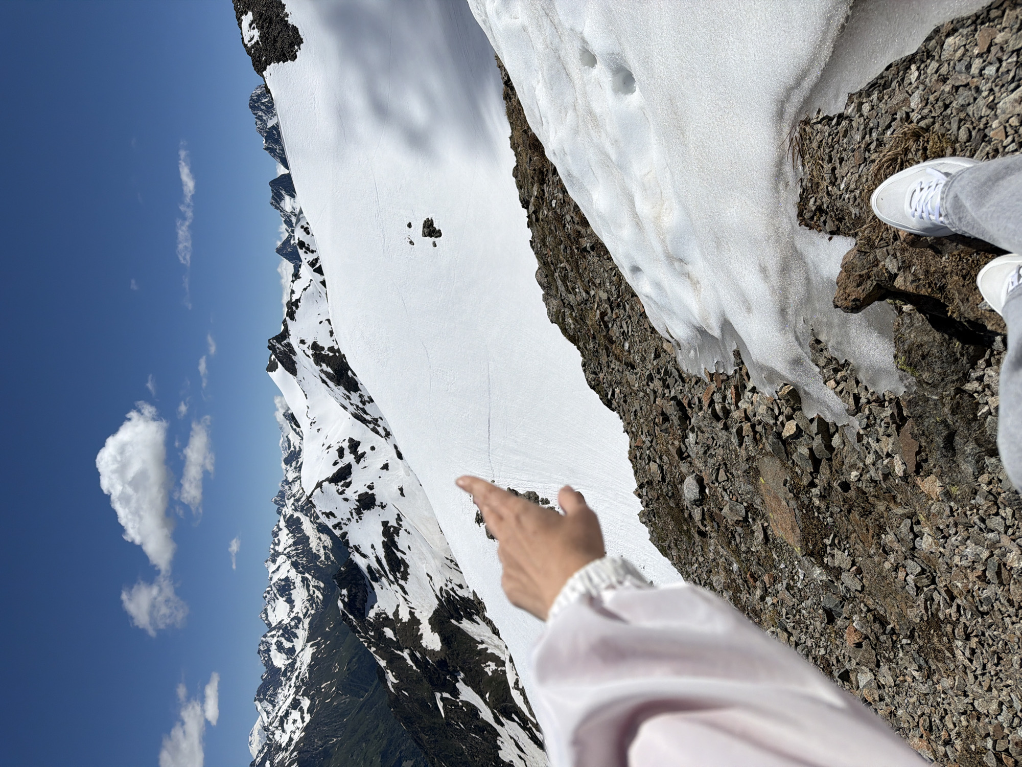 A person's hand, pointing towards a snow-covered mountain range, is visible in the foreground of the image. The person is wearing a light-colored jacket. The background features a vast expanse of snow-capped mountains under a clear blue sky with scattered clouds. The terrain in the foreground consists of rocks and patches of snow. The location is the city of Verkhne-Teberdinskoye rural settlement, Russia.