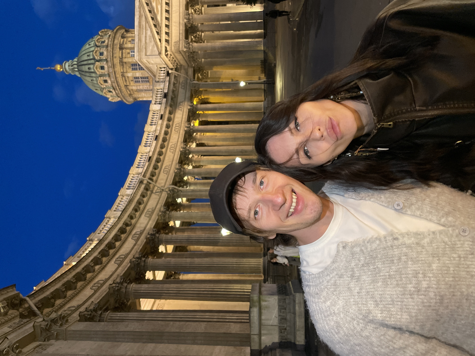 In Saint Petersburg, Russia, a couple takes a selfie at night in front of Kazan Cathedral. The man, wearing a light gray knitted cardigan over a white t-shirt and a black baseball cap, smiles widely. The woman, with long dark hair and wearing a dark brown leather jacket, pouts her lips. Behind them, the impressive neoclassical facade of Kazan Cathedral is illuminated, its grand colonnade and dome silhouetted against a dark blue twilight sky. Several streetlights dot the plaza in front of the cathedral, and a few distant figures can be seen walking. The lighting suggests it is late evening.