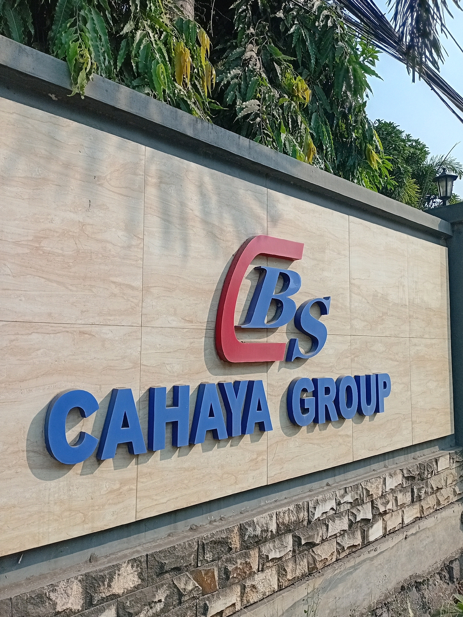 The image shows the exterior signage of "CBS CAHAYA GROUP" located in Cilegon, Indonesia. The sign is mounted on a beige tiled wall. The logo, a red stylized "C" encompassing blue letters "BS", is prominently displayed above the company name "CAHAYA GROUP" in large blue 3D letters. The building is framed by lush green foliage at the top and a stone-clad base at the bottom. The sky is visible in the upper right corner, and electrical wires are strung across the top. The lighting suggests it is daytime, with shadows cast by the signage.