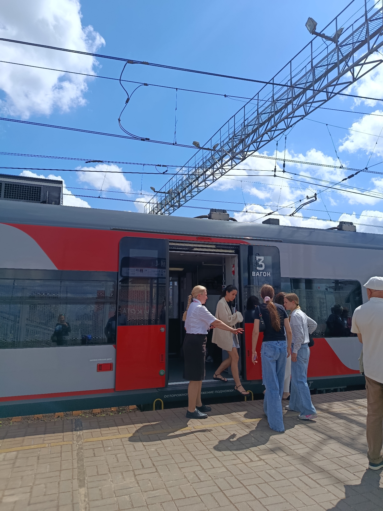 At a train station in Orekhovo-Zuyevo, Russia, a train with red and grey cars is stopped at a platform under a bright blue sky with scattered clouds. Overhead, a complex network of electrical wires and support structures for the train's power system is visible. Several people are standing on the platform near the open doors of the train. A woman in a white shirt and dark skirt, appearing to be a train attendant, is gesturing towards the train entrance. A group of passengers, including several young women, are either boarding or disembarking. The platform is paved with light-colored bricks, and a yellow line marks the edge of the platform. Reflections of people and the train interior are visible in the train windows. The overall scene captures a moment of passenger activity at a Russian train station.