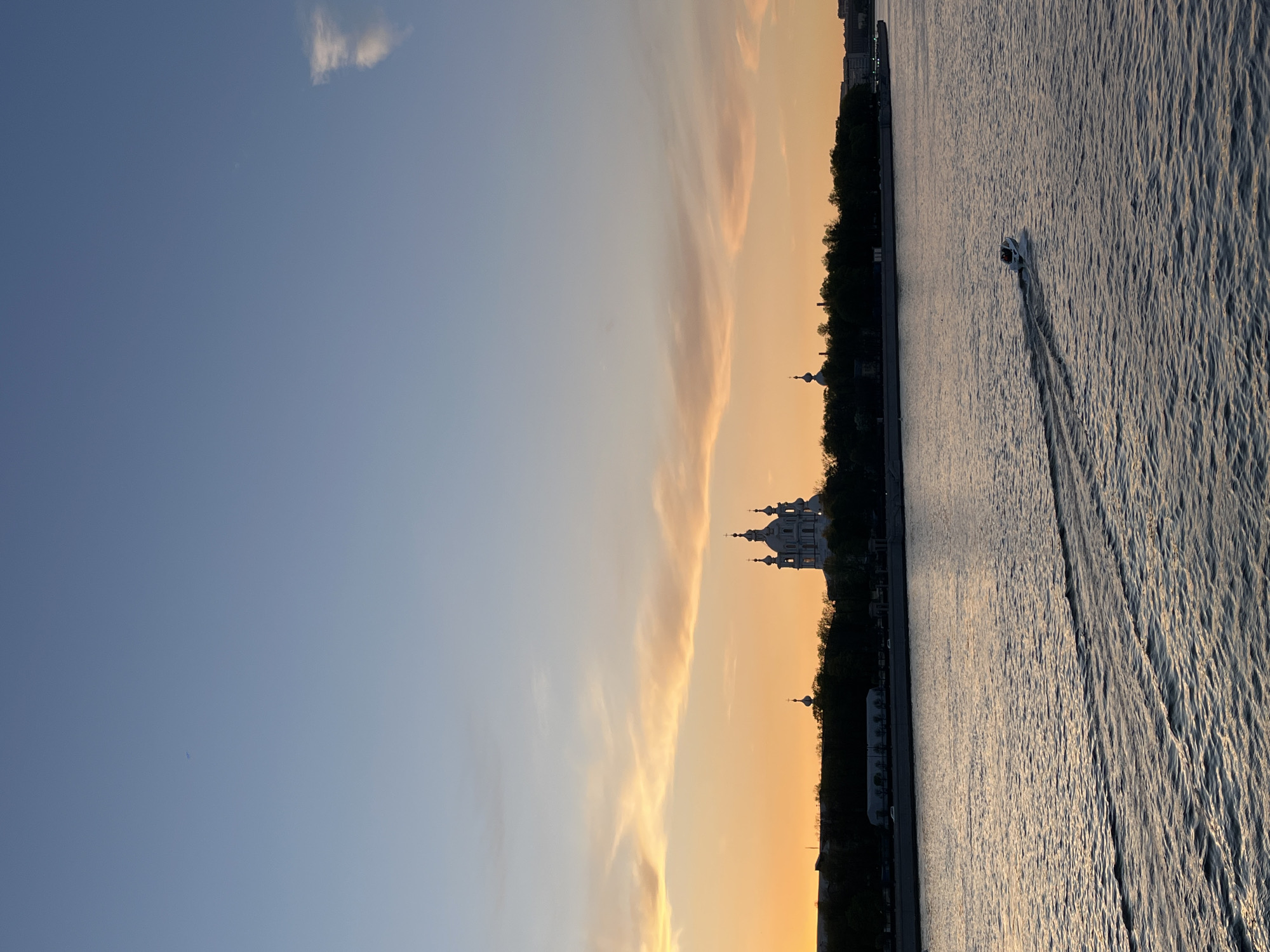 The image depicts a riverside view at sunset in Saint Petersburg, Russia. The Smolny Cathedral is prominently silhouetted against the setting sun. A small motorboat is moving on the water, leaving a wake behind it. The sky transitions from blue to golden hues, with wisps of clouds visible. Trees line the embankment along with building silhouettes.