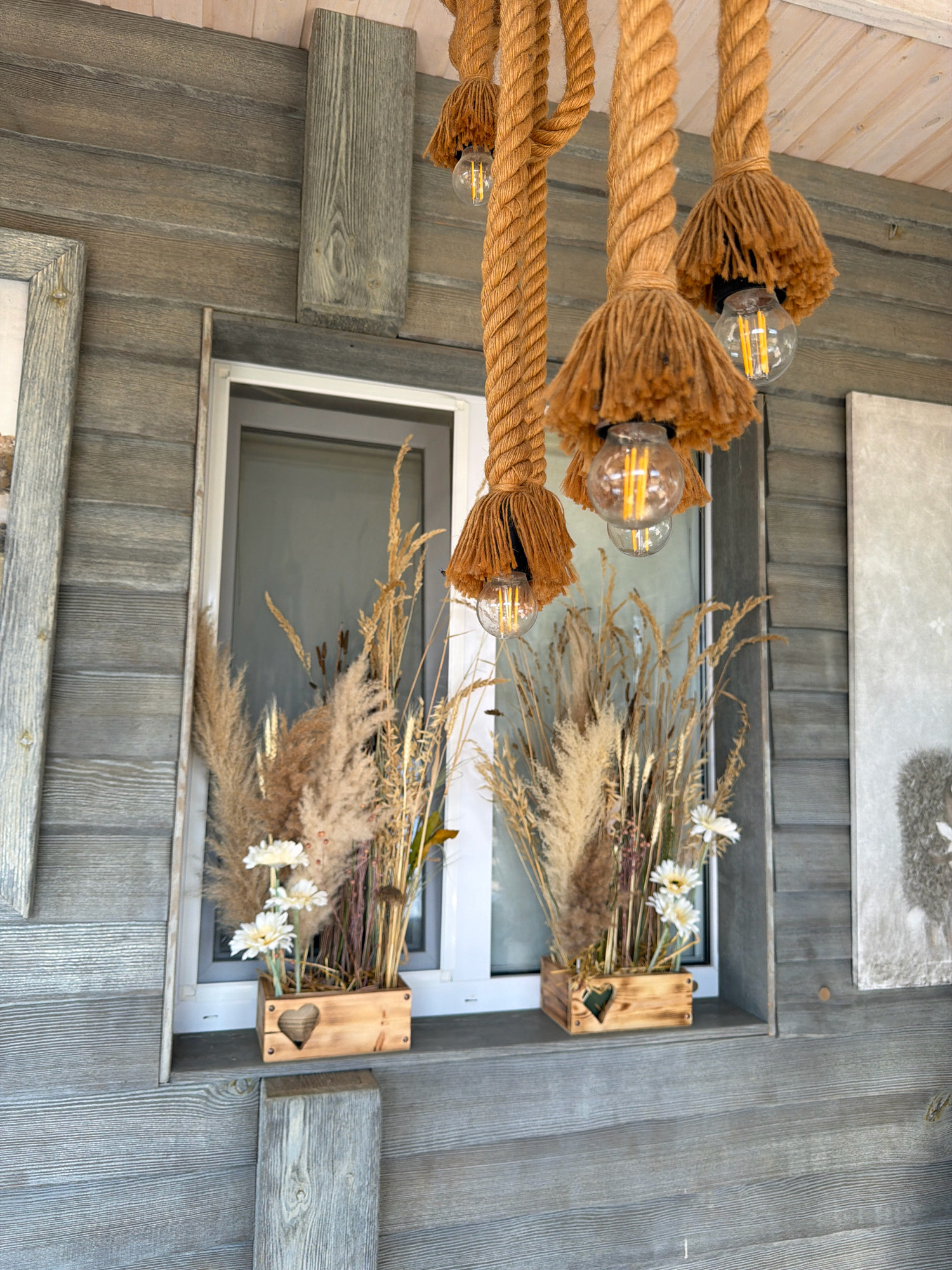 The image shows a close-up of a wooden-clad exterior wall with a window. Three decorative rope pendant lights with exposed bulbs and tassel-like shades hang from above. Below the window, on the windowsill, are two wooden planters filled with dried grasses and two white flowers each. The planters have a cutout heart shape on the front. The wall is constructed of horizontally laid wooden planks with a weathered grey finish. A portion of a framed artwork or picture is visible on the left side of the image, and another on the right side. The overall setting appears to be an outdoor area, possibly a cafe, restaurant, or porch, in the village of Perovske, Ukraine.