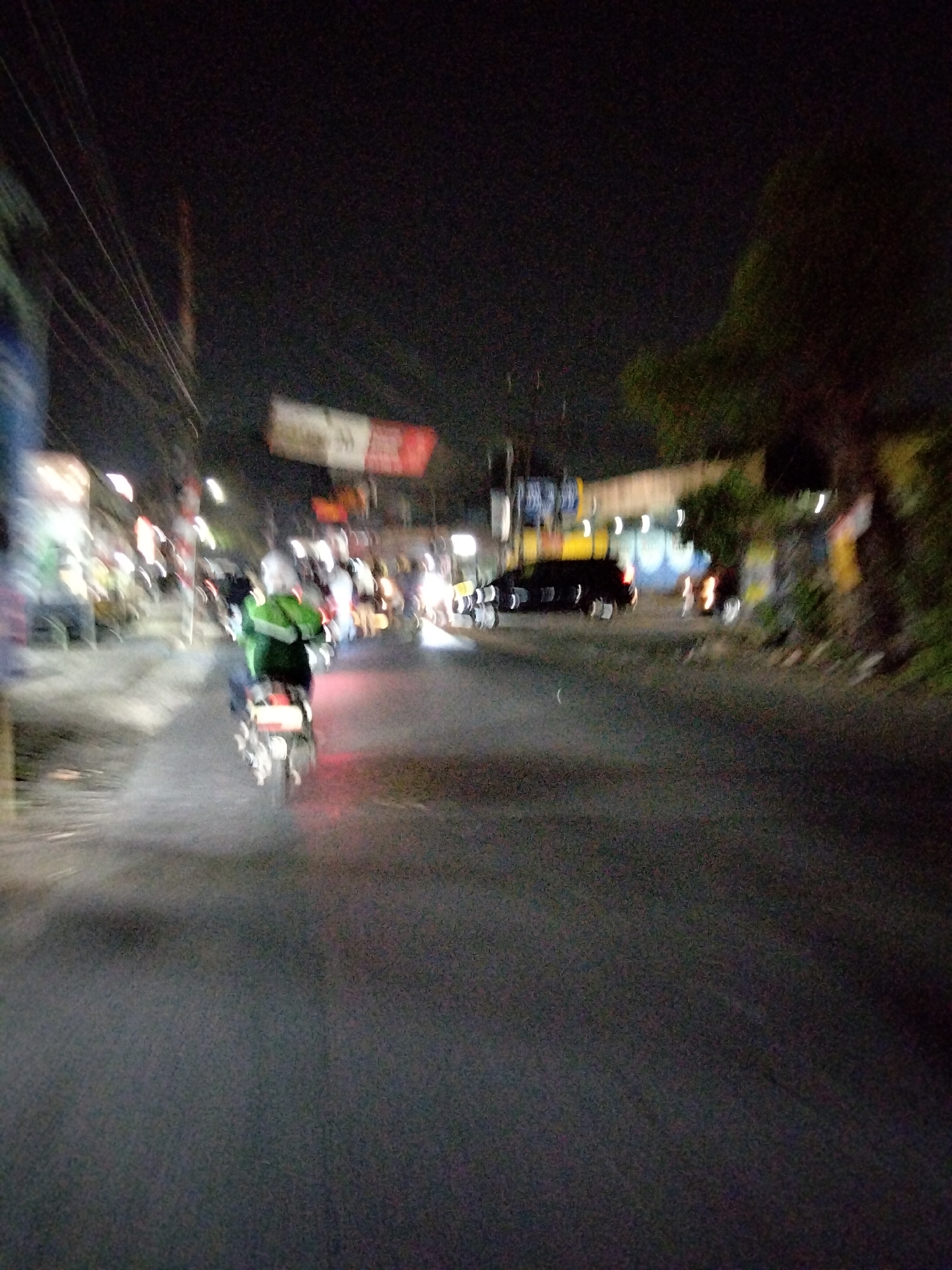 The image depicts a street scene at night in South Tangerang, Indonesia. The camera appears to be moving, as indicated by motion blur. In the foreground, a person is riding a motorcycle, wearing a green jacket and black pants. The motorcycle's taillight is illuminated red. To the left, there are blurred figures of people and possibly stalls or shops, with bright lights creating streaks. In the background, a dark-colored car is visible on the road, with its taillights also illuminated. Trees and vegetation can be seen on the right side of the street. Overhead, utility wires are present. A sign with red and white elements is blurred in the upper center of the image. The overall atmosphere is one of a busy, illuminated street at night.