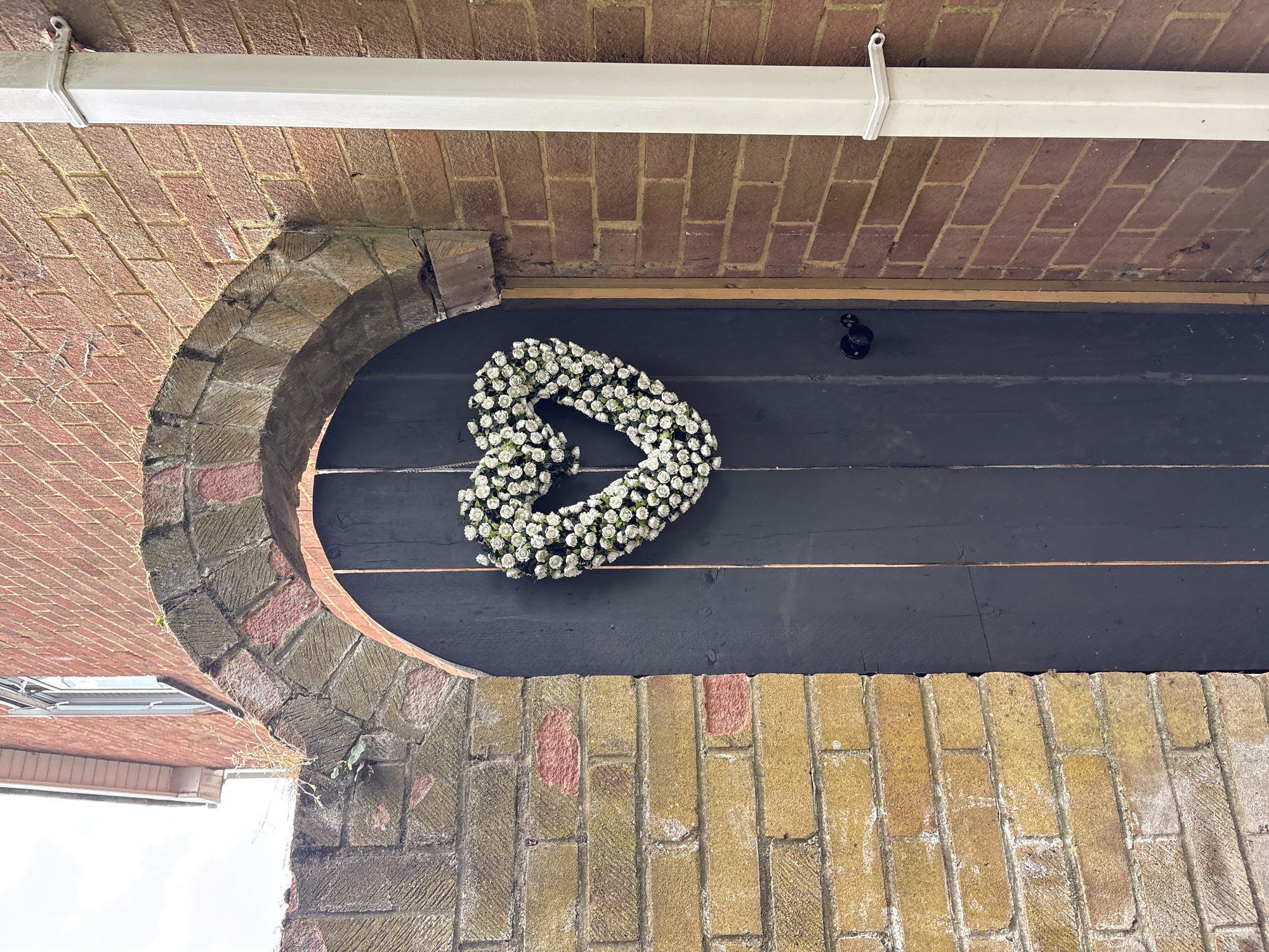 The image shows a dark wooden door with a heart-shaped wreath of white flowers hanging on it. The door is set within an arched opening made of rough-hewn stone and brick. To the right of the door, a white downpipe runs vertically along a red brick wall. The scene appears to be an exterior shot, possibly the entrance to a property. The architecture suggests an older building. The image is taken from a slightly low angle, looking up towards the archway and the top of the door. There are no people visible in the image. The lighting is overcast, indicating a cloudy day. The environment appears to be domestic or residential, with the brickwork and architectural style characteristic of many properties in Chelmsford, United Kingdom.