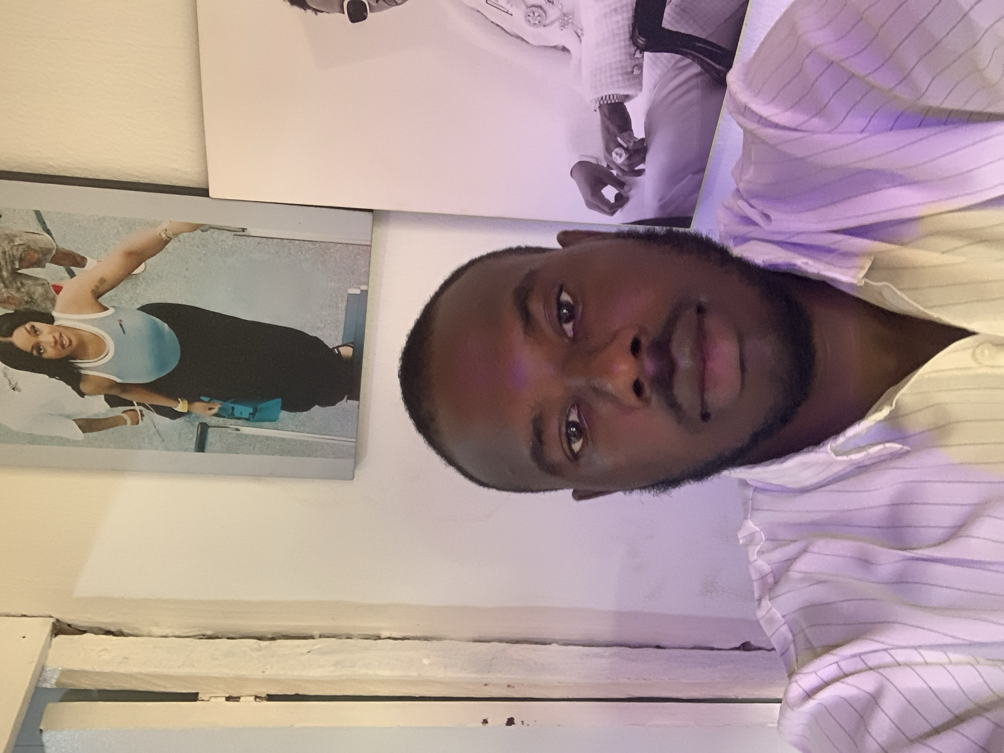 A close-up, tilted portrait captures a Black man with a trimmed beard wearing a white button-down shirt with light purple pinstripes. Behind him, two framed photographs hang on a white wall. The photo on the upper right depicts a person in white clothing adorned with jewelry, while the photo on the left shows a woman in exercise attire. The location in Jos, Nigeria is not discernible in the provided image.