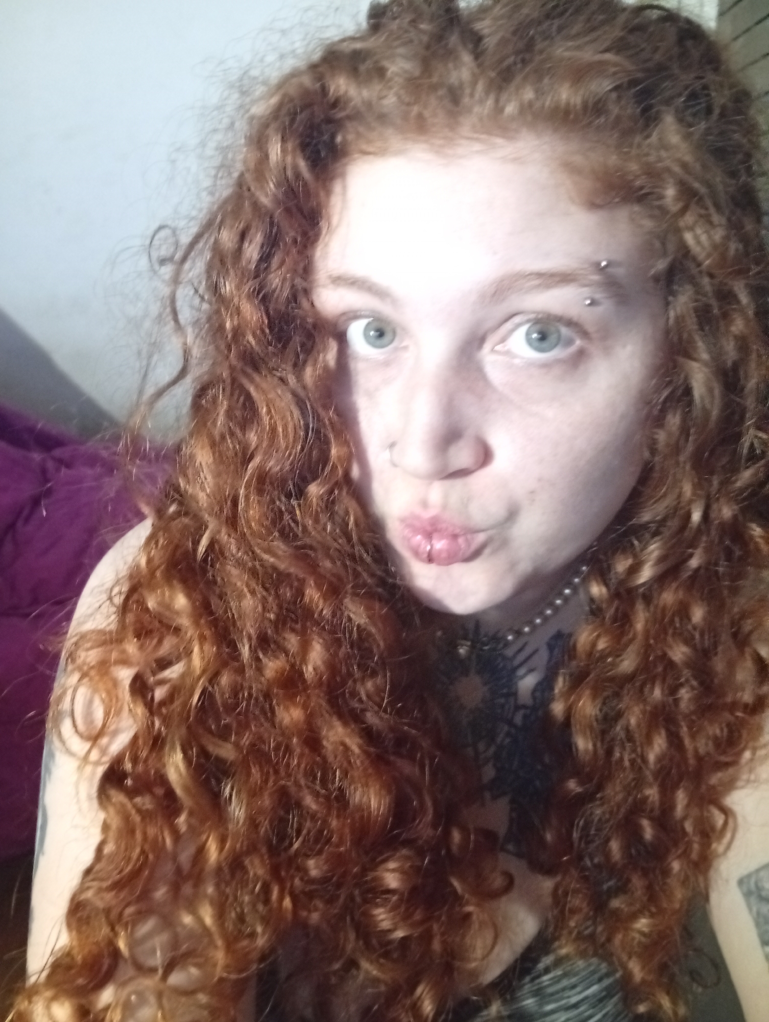 A selfie of a pale-skinned woman with long, curly red hair. She has light blue eyes, a nose ring, and an eyebrow piercing. She is making a kissing face. A pearl necklace and tattoos are visible on her chest and arms. A purple object is behind her, out of focus. The location is not identifiable as Buenos Aires, Argentina.