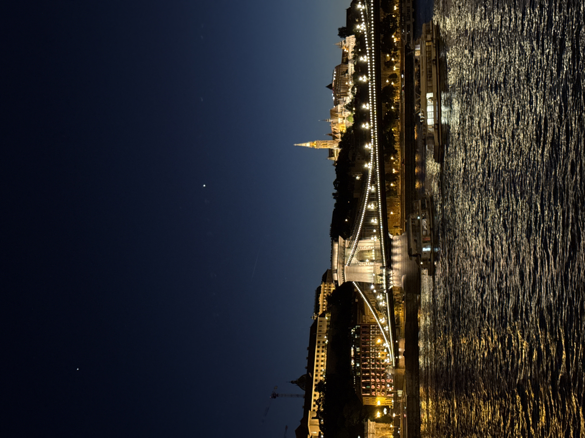 The image captures a nighttime view of Budapest, Hungary, featuring the Széchenyi Chain Bridge illuminated with lights. The Danube River reflects the bridge's lights, creating a shimmering effect. In the background, the Buda Castle and Matthias Church are visible, also illuminated. The sky is dark, suggesting twilight or early evening.