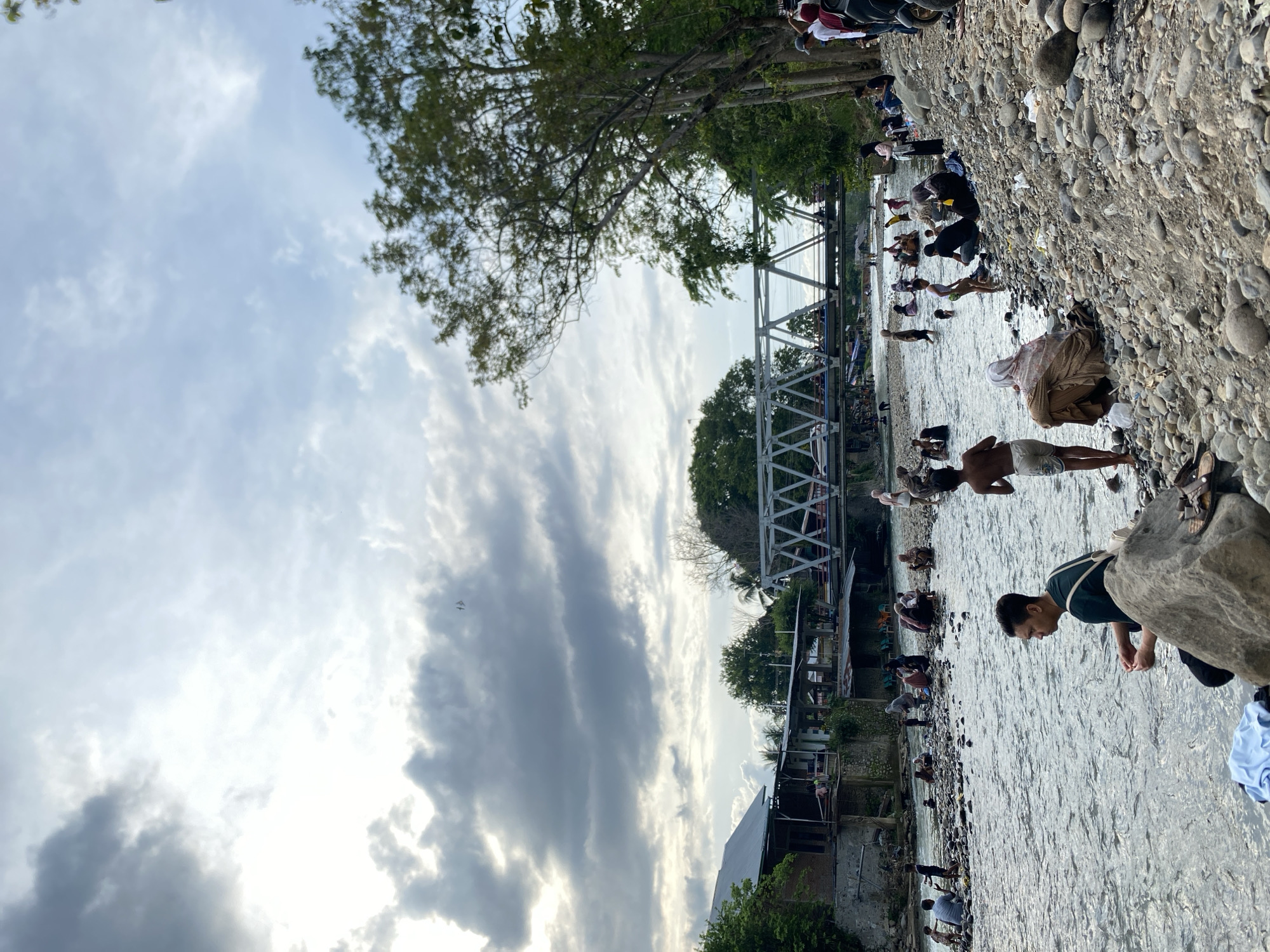 People are gathered in and along the banks of a river in Meurah, Indonesia. A pedestrian bridge crosses the river. The riverbank is composed of smooth stones. People are engaging in various activities in the river, including bathing and washing clothing. Houses and buildings are visible on one bank, amidst trees and vegetation. The sky is cloudy.