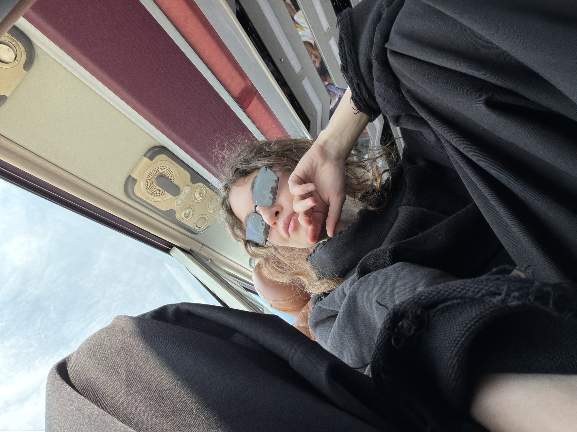 A low-angle close-up photograph captures a person, likely a young woman, inside what appears to be a bus or train in Hải Phòng, Vietnam. The subject is seen from a perspective looking upwards, suggesting they are reclining or positioned low in their seat. She has curly brown hair and wears dark sunglasses, which reflect the bright, cloudy sky outside. Her right hand is raised to her face, partially obscuring her mouth and chin.
She is dressed in loose-fitting black clothing, possibly a hoodie or top and trousers, with visible distressed or frayed edges on the sleeves. Behind her head, a light brown or tan object, possibly a seat headrest, is visible. The interior of the vehicle features overhead panels with integrated lights and air vents, colored in muted tones of cream and burgundy. A large window on the left side of the frame shows a bright, partially cloudy daytime sky. The overall impression is a casual, relaxed moment of travel. No visible text is present in the image.