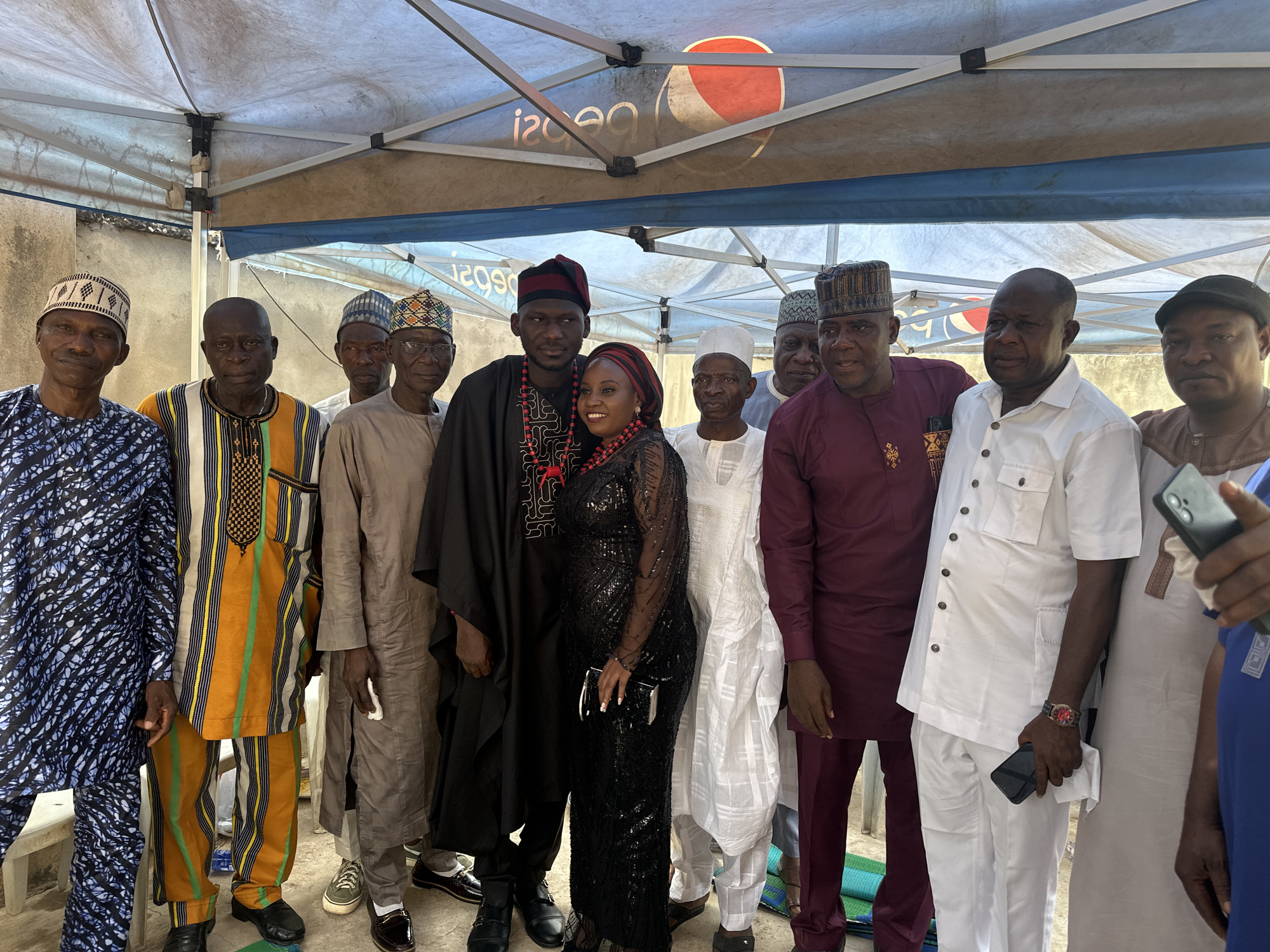 The image displays a group of ten adults, comprising nine men and one woman, posing together under a temporary canopy structure. The setting is outdoors, likely during daylight hours, suggested by the bright lighting and the overhead canopy providing shade. The attire of most individuals suggests a cultural gathering or celebration, possibly in an African context, with many wearing traditional clothing.
At the center of the group, a man and a woman appear to be the focal point. The man wears a black traditional tunic adorned with red and black beaded necklaces and a matching cap. Beside him, the woman smiles broadly, dressed in a long, black sequined gown with long sleeves, a red and black patterned headscarf, and coordinating beaded jewelry. She holds a small black clutch bag.
The other men in the group are dressed in a variety of traditional garments. These include a man in a blue and white patterned outfit, another in a vibrant yellow, orange, green, and black striped dashiki, and several men in different colored long tunics (light brown, white, maroon) often paired with patterned caps. One man wears a white short-sleeved button-down shirt and white trousers.
The overhead canopy, supported by metal frames, shows a reversed "Pepsi" logo ("isdeP") and the numbers "120" (also reversed) printed on its underside. The ground beneath them is a mix of concrete and dirt, with a blue and green mat visible towards the right. The overall atmosphere appears pleasant and celebratory.
