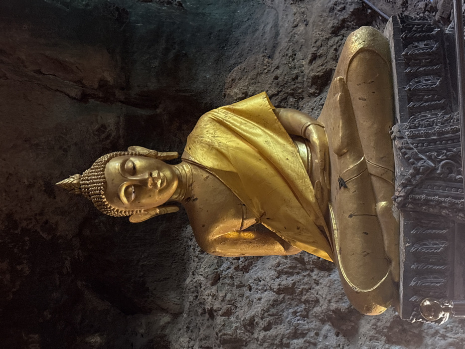 A golden Buddha statue is centrally positioned within a cave in Ban Hin Sam Kon, Thailand. The statue is depicted seated in a cross-legged meditative posture, with its hands resting in its lap. Its surface is golden-colored, featuring a flame-like ushnisha, tightly curled hair, closed eyes, and elongated earlobes. A draped, shiny golden fabric covers the statue's left shoulder and torso. Minor dark smudges and surface wear are visible on the statue, particularly on its legs. It rests upon a multi-tiered, dark-colored base adorned with intricate, dark relief carvings. The background consists of dark, textured rock walls, indicating a natural cave environment, illuminated by an unseen light source that highlights the statue.