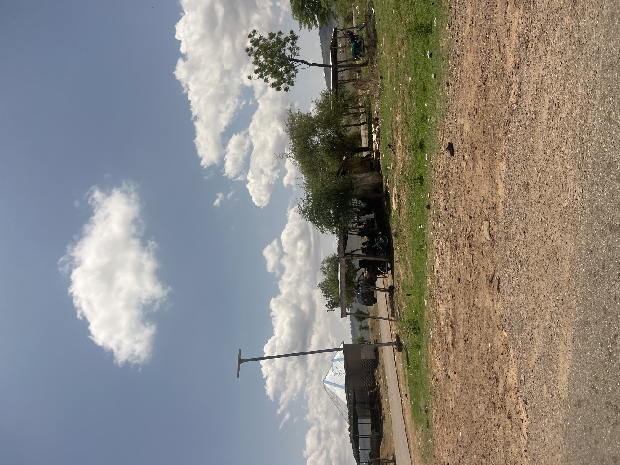 This image depicts a semi-rural roadside scene in Chum, Nigeria, under a bright, partly cloudy sky. The light blue sky is populated with numerous white cumulus clouds of varying sizes.
In the foreground, an asphalt road runs horizontally across the bottom. Immediately above it, a wide stretch of reddish-brown dirt and gravel ground extends into the mid-ground, interspersed with patches of green grass and scattered small debris.
Mid-ground elements include:
On the far left, a structure with a distinctive conical or pyramidal roof, featuring alternating light blue and white stripes. Adjacent to it, a smaller, darker structure is visible under a flat roof.
Towards the center-left, a tall, slender dark gray pole, possibly a solar street light, stands upright beside a paved road that curves slightly to the right.
Further right, a series of rudimentary shelters or kiosks are constructed with wooden posts supporting flat, dark roofs. Under one of these shelters, a dark-colored car is partially parked, and several individuals are visible, appearing to be gathered or sitting. Another green motorcycle is parked under a separate shelter to the right.
Numerous green trees and bushes are scattered throughout the mid-ground, some dense, others more sparse, with one prominent tree featuring a tall trunk and rounded canopy on the far right.
In the background, gently rolling hills or low mountains covered in light green vegetation are visible beneath the horizon.