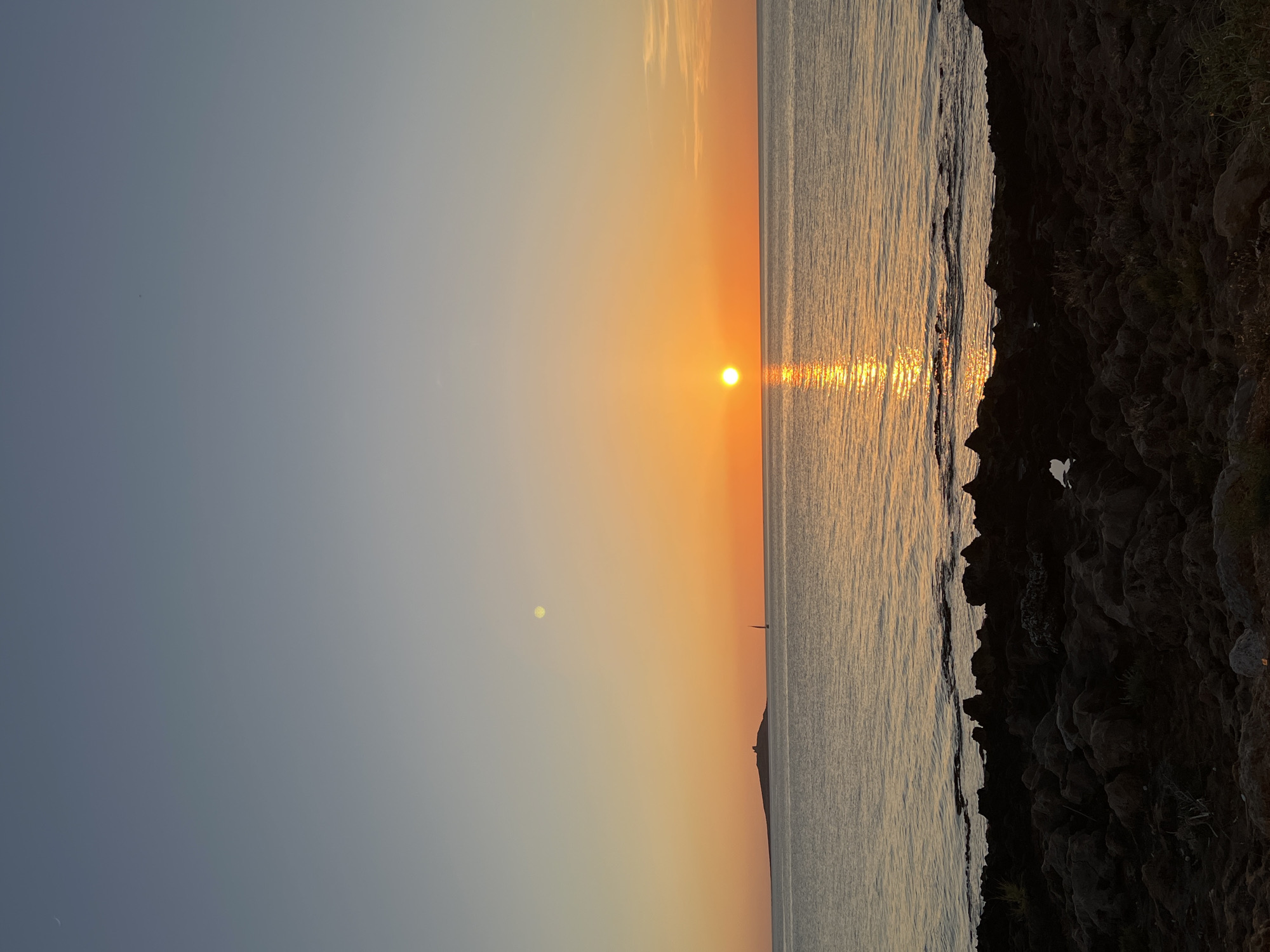 The image depicts a sunset over the sea, with a rocky shoreline in the foreground. The sun is low on the horizon, casting a warm orange glow across the sky and water. In the distance, a small island with a building on it is visible, along with a sailboat. The sky transitions from a deep blue at the top to a lighter orange near the horizon. The rocks in the foreground are dark and jagged, with some sparse vegetation. The water is calm, with gentle ripples reflecting the sunlight. The scene appears to be a coastal area, possibly outside of the city center of Palermo, Italy, given the natural landscape. There are no people or animals clearly visible in the frame.