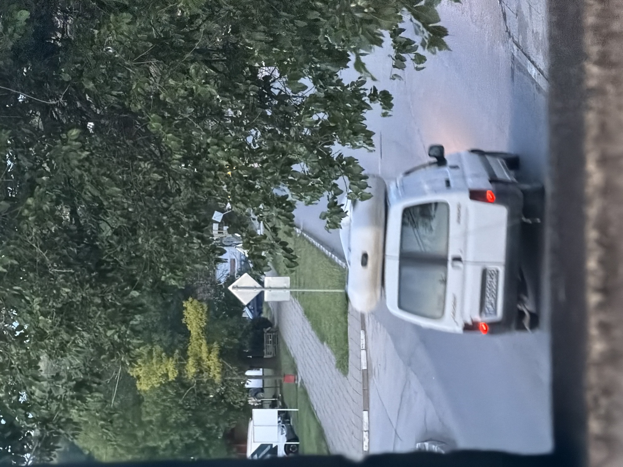 The image, taken from an elevated perspective and oriented vertically, captures a street scene in Iskar, Bulgaria, during what appears to be late afternoon or early evening under a diffused, possibly overcast sky.
The primary subject is a white panel van, possibly a Citroën Berlingo or similar model, driving away from the viewer on a paved road. Its red taillights are visible, and the Bulgarian license plate "CB 5859 PH" is clearly legible on its rear. The "CB" prefix indicates Sofia, consistent with Iskar being a town in Sofia Province.
To the left, dense green foliage from numerous trees and bushes dominates a significant portion of the frame, creating a natural canopy over parts of the street and sidewalk. Some patches of yellowish-green foliage are also visible further in the background. A gray sidewalk runs parallel to the road, separated by a concrete curb and a strip of grass. A diamond-shaped road sign, too blurry for specific text but clearly identifiable by its form, stands on a pole next to the sidewalk.
Other vehicles, including another white van and a darker car, are partially visible parked or moving further down the street behind the trees. The scene is framed on the right by a dark, textured wall or window frame, suggesting the photo was taken from inside a building or from a balcony. There are no people visible, and no specific activity beyond the van driving is clearly depicted. The overall impression is that of a quiet, tree-lined suburban or semi-urban street.