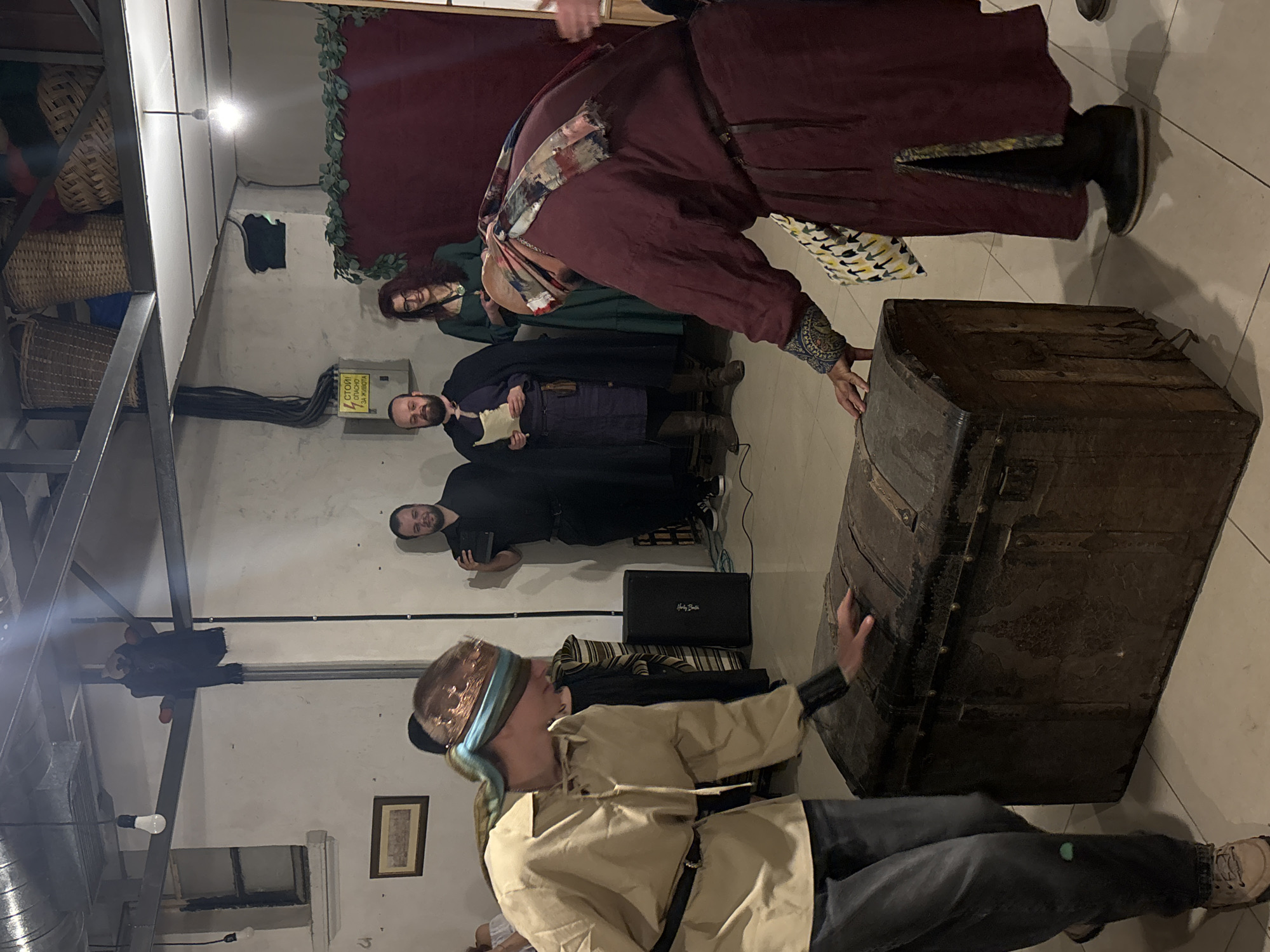 Captured indoors, the scene showcases several individuals seemingly involved in a play or performance. In the foreground, a person in a beige garment, blindfolded with a teal and copper cloth, kneels near an old, dark-colored chest. Behind them and to the left, additional individuals stand or kneel, adorned in what appear to be theatrical costumes. One figure, cloaked in a maroon robe with intricate sleeve detailing, leans over the chest. The backdrop includes a wall partially covered with a red curtain and artificial greenery. There's electrical wiring exposed on the wall near a rectangular sign. A speaker is placed near the individuals in costume.
Based on the architecture and setting, this event may be taking place in an indoor space in Varna, Bulgaria, although there are no definitive landmarks visible in the image to confirm this location.