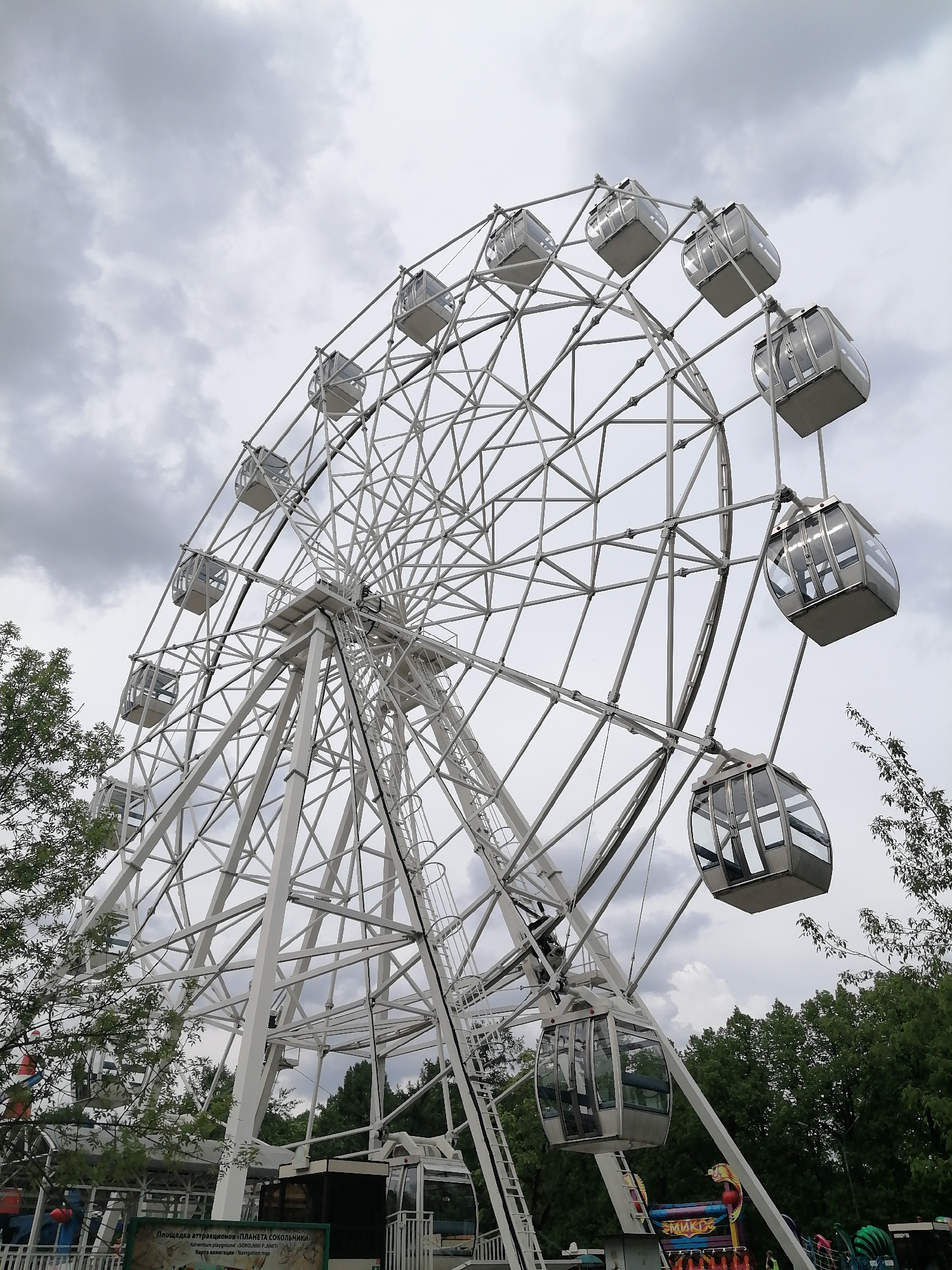 The image depicts a large white Ferris wheel at an amusement park. The wheel has several enclosed gondolas. The sky is cloudy. The park is located in Sokolniki Park in Moscow, Russia, indicated by the sign "SOKOLNIKI PLANET". In the foreground, green trees and other park attractions are visible.