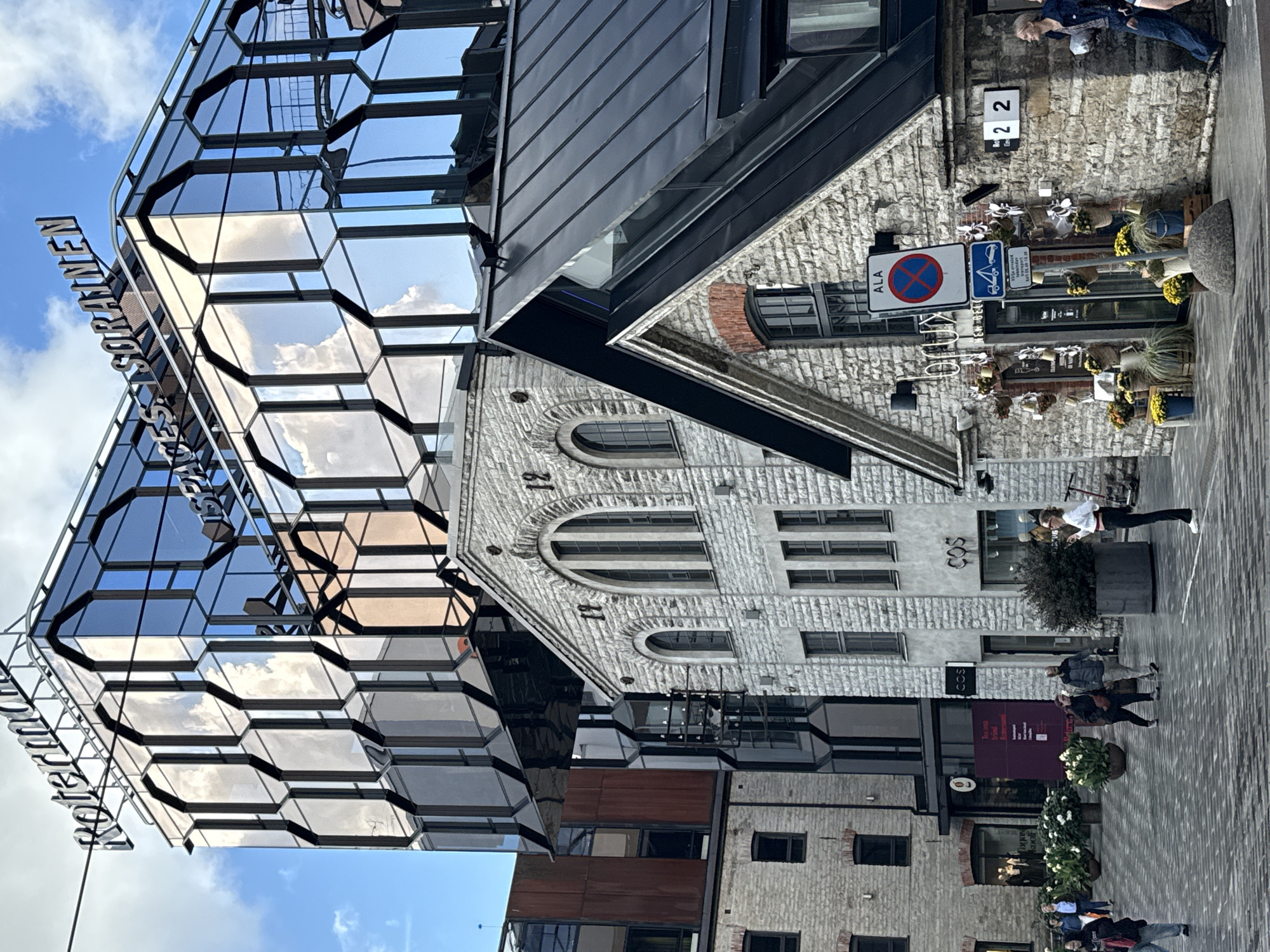 The image depicts a street scene in the Rotermann Quarter of Tallinn, Estonia. In the background, a modern glass building with a distinctive geometric facade, bearing the text "Rotermann" and "SPACES SORAINEN," stands out against a partly cloudy sky. In the mid-ground, two older stone buildings are visible. The building on the left has arched windows and a textured stone facade. The building on the right, with a dark metal roof, has a stone facade and features a storefront with potted plants and decorative flowers. A "2 2" number sign is affixed to its wall. In the foreground, people are walking on a paved street. A woman in a white shirt and black pants stands to the right of the stone building, facing left. The scene captures the architectural blend of old and new that characterizes the Rotermann Quarter.