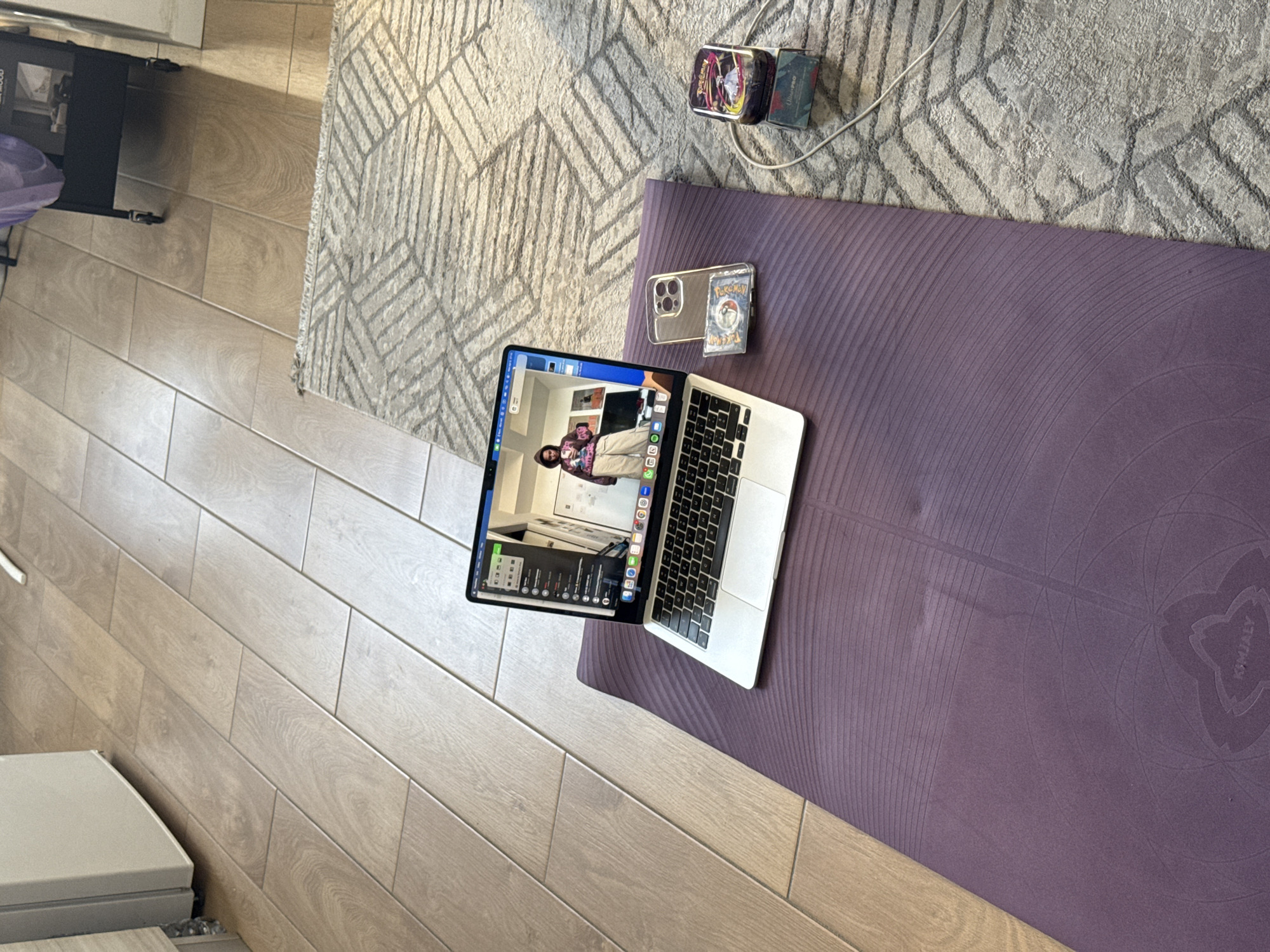 The scene shows a purple yoga mat laid out on a light-colored wooden floor. A silver laptop is placed on the mat, displaying a video call. A smartphone in a clear case with a Pokemon card attached to it rests beside the laptop on the mat. A Pokemon card box is connected to the charger of the phone, placed on a grey patterned rug. The location is not identifiable as Milan, Italy from the scene.