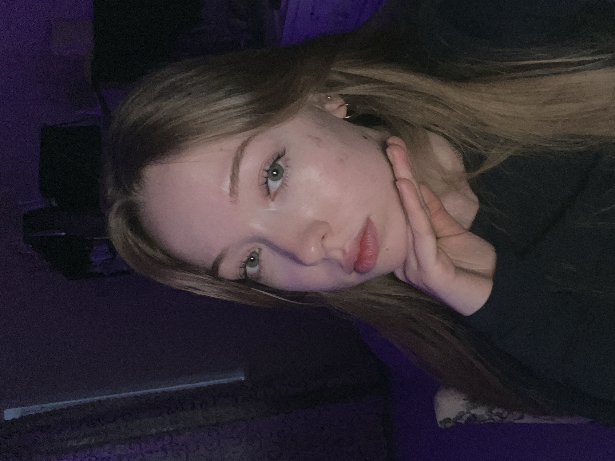 The image is a close-up selfie of a woman with light brown hair and green eyes, who appears to be resting her cheek on her hand. She is wearing a dark-colored top. The background is dark and purple-toned, suggesting low or artificial lighting. No information indicates that this image was taken in Chaikovsky, Russia, or any other location.