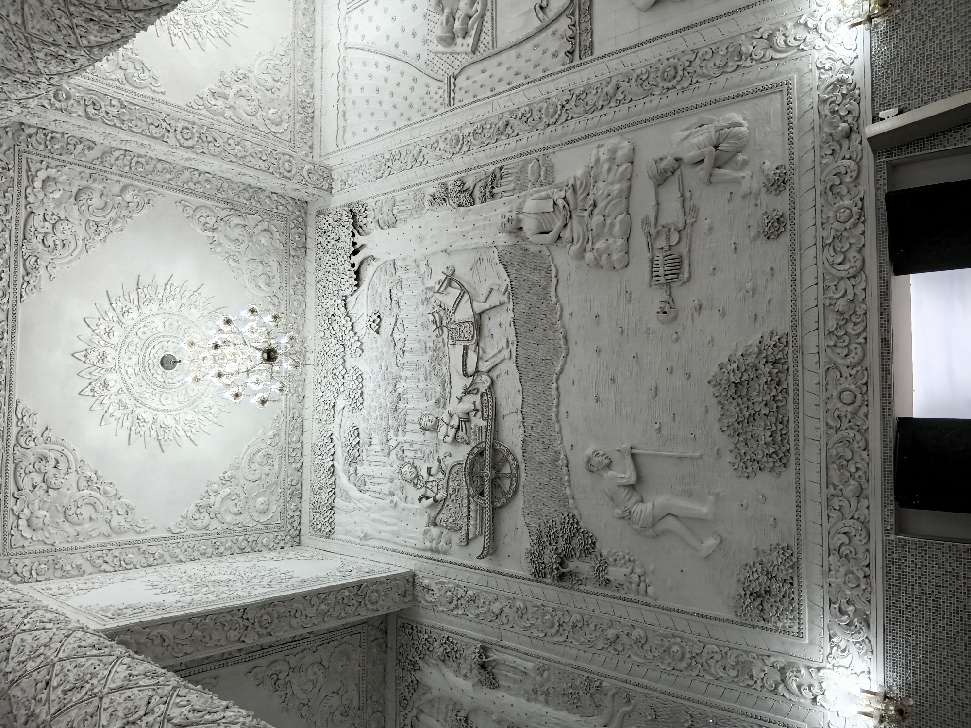 The image depicts an interior scene of a temple, likely in Ban Lao Phatthana, Thailand. The dominant visual element is intricate white relief art covering the walls and ceiling. The relief art portrays various scenes, including figures in a chariot pulled by a horse, a seated Buddha figure under a tree, a person with a staff, and a skeleton with a kneeling figure. The ceiling is adorned with ornate patterns and features a chandelier. The overall aesthetic is highly decorative and detailed, with a monochromatic color scheme of white. The lighting suggests it is an indoor environment, possibly a place of worship or cultural significance.