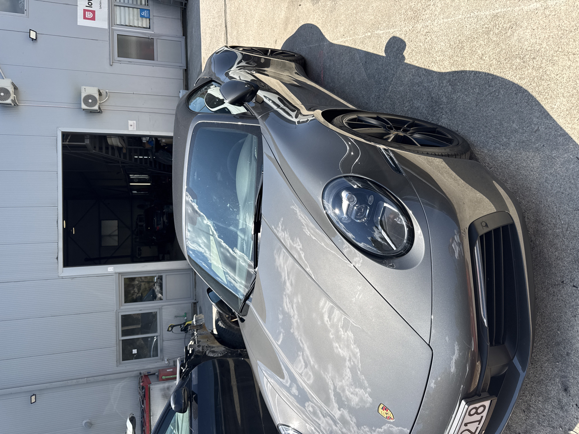 The image depicts a gray Porsche convertible parked on a concrete surface outside a modern industrial building, possibly a car service or repair shop. The building features a large, open bay with a metal staircase visible inside, and a gray corrugated metal facade. There are also several windows and doors on the building's exterior. A black car, partially disassembled, is parked to the left of the Porsche. The scene is illuminated by bright sunlight, casting distinct shadows on the ground. The sky, with scattered clouds, is reflected on the car's polished surface. A license plate is visible on the front of the Porsche, partially obscured. The location is identified as Dub, Montenegro, suggesting this is an outdoor area associated with businesses in that region. There are no people visible in the image.
