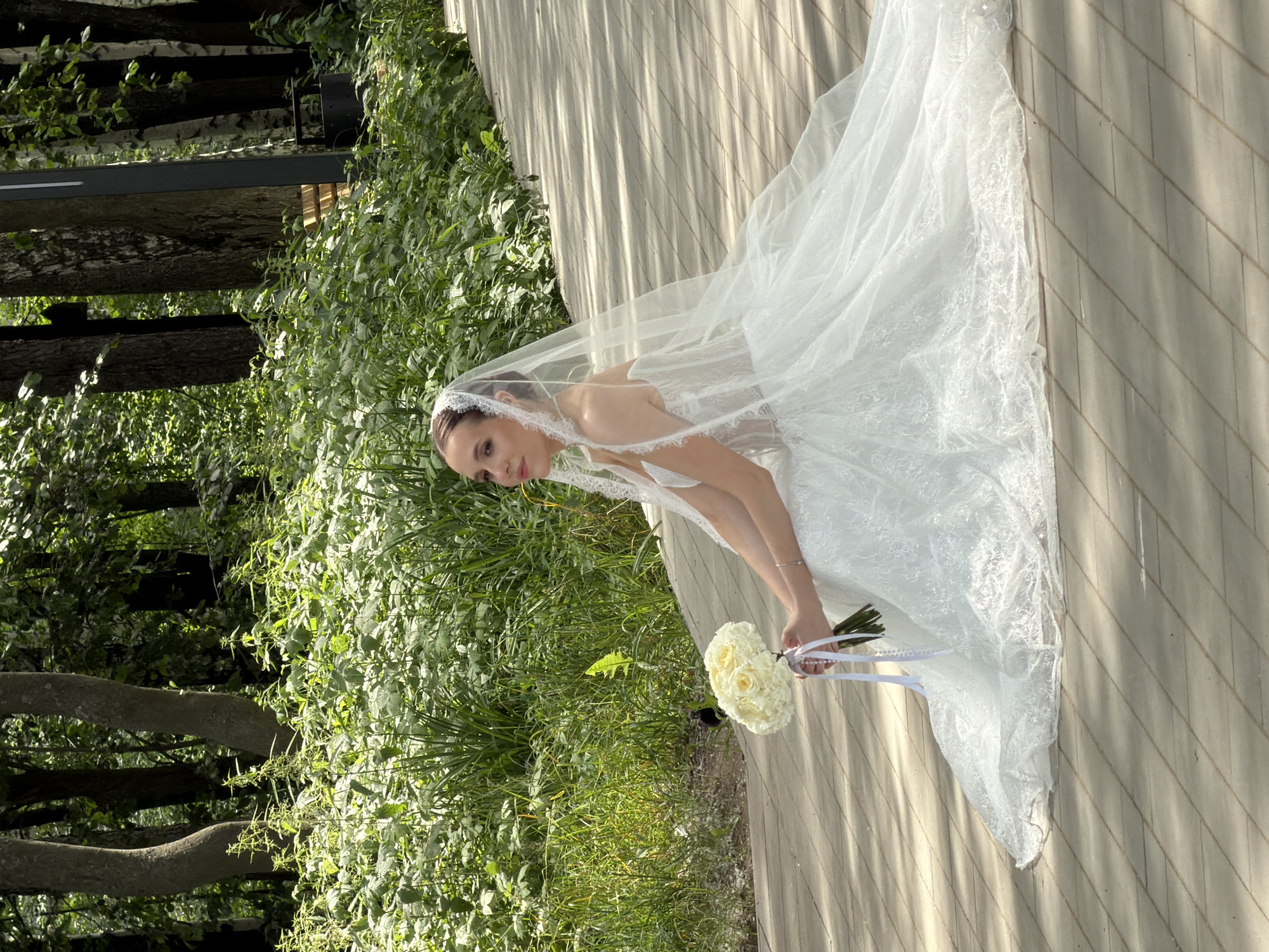 This media file shows a bride in a white wedding dress and veil. She is holding a bouquet of white roses with white ribbon accents. The bride is looking down and to her left, with a soft expression on her face. The scene appears to be outdoors, with lush green foliage on the left side of the frame and a wooden deck or walkway on the right. The lighting suggests it is daytime, possibly late afternoon given the long shadows cast by the sun on the wooden surface. The overall mood is serene and romantic, fitting for a wedding. The location context is provided as Chaikovsky, Russia. No visible text is present in the image.
