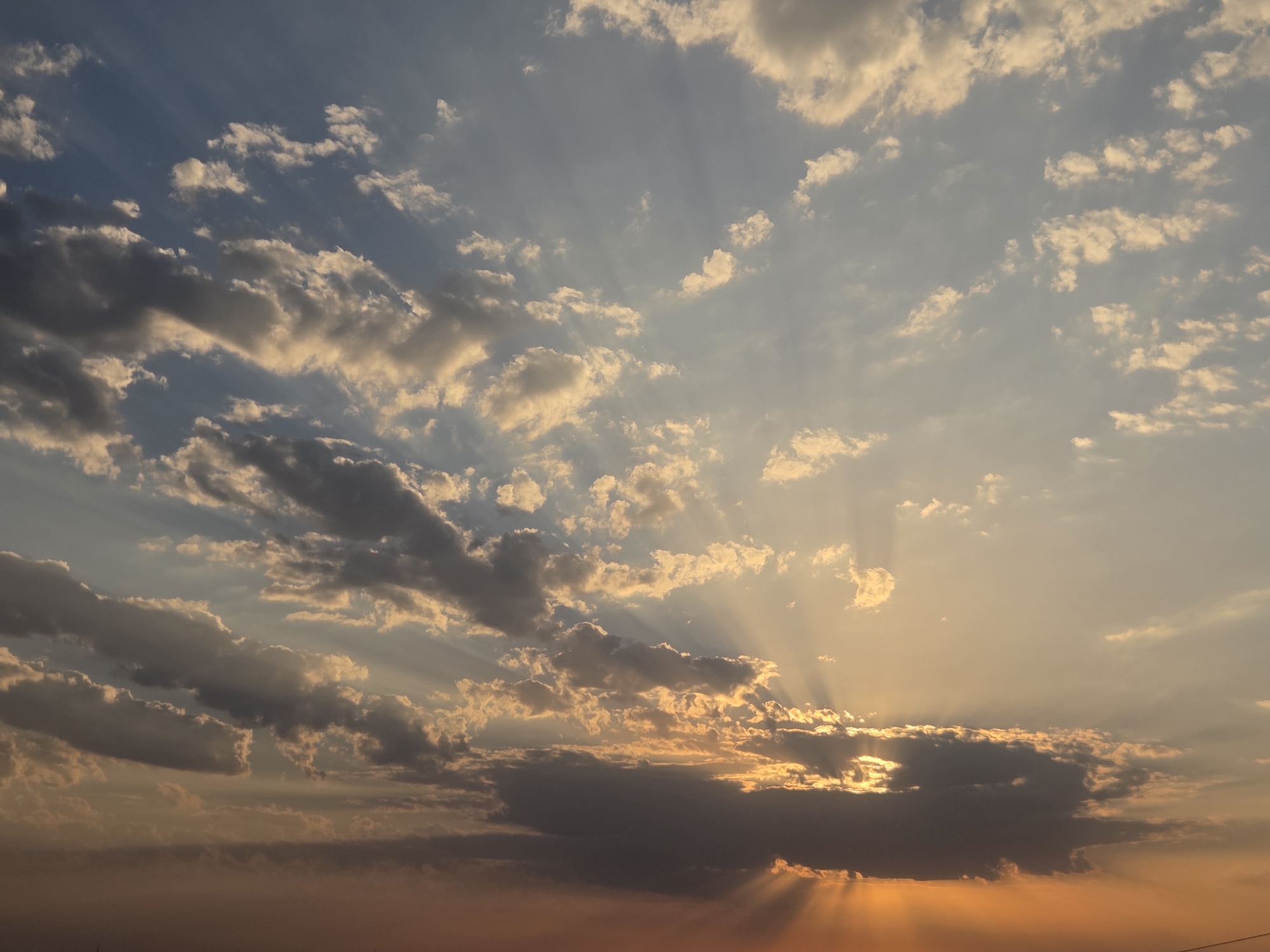 The image depicts a sky filled with a dramatic interplay of sunlight and clouds, characteristic of a sunset or sunrise. The sun's rays are visible, piercing through gaps in the clouds, creating crepuscular rays that fan out across the sky. The clouds themselves vary in appearance, with some appearing dark and heavy, suggesting lower altitudes or denser formations, while others are lighter and more scattered. The lower portion of the sky exhibits warm, orangey-yellow hues, transitioning to cooler blues and grays higher up, indicating the direction of the setting or rising sun. The scene is entirely focused on the sky, with no visible land features or man-made structures, therefore, it is not possible to determine the specific location within the city of Қарасу, Kazakhstan, or to identify any people, subjects, objects, or actions. The environment is solely the celestial expanse.