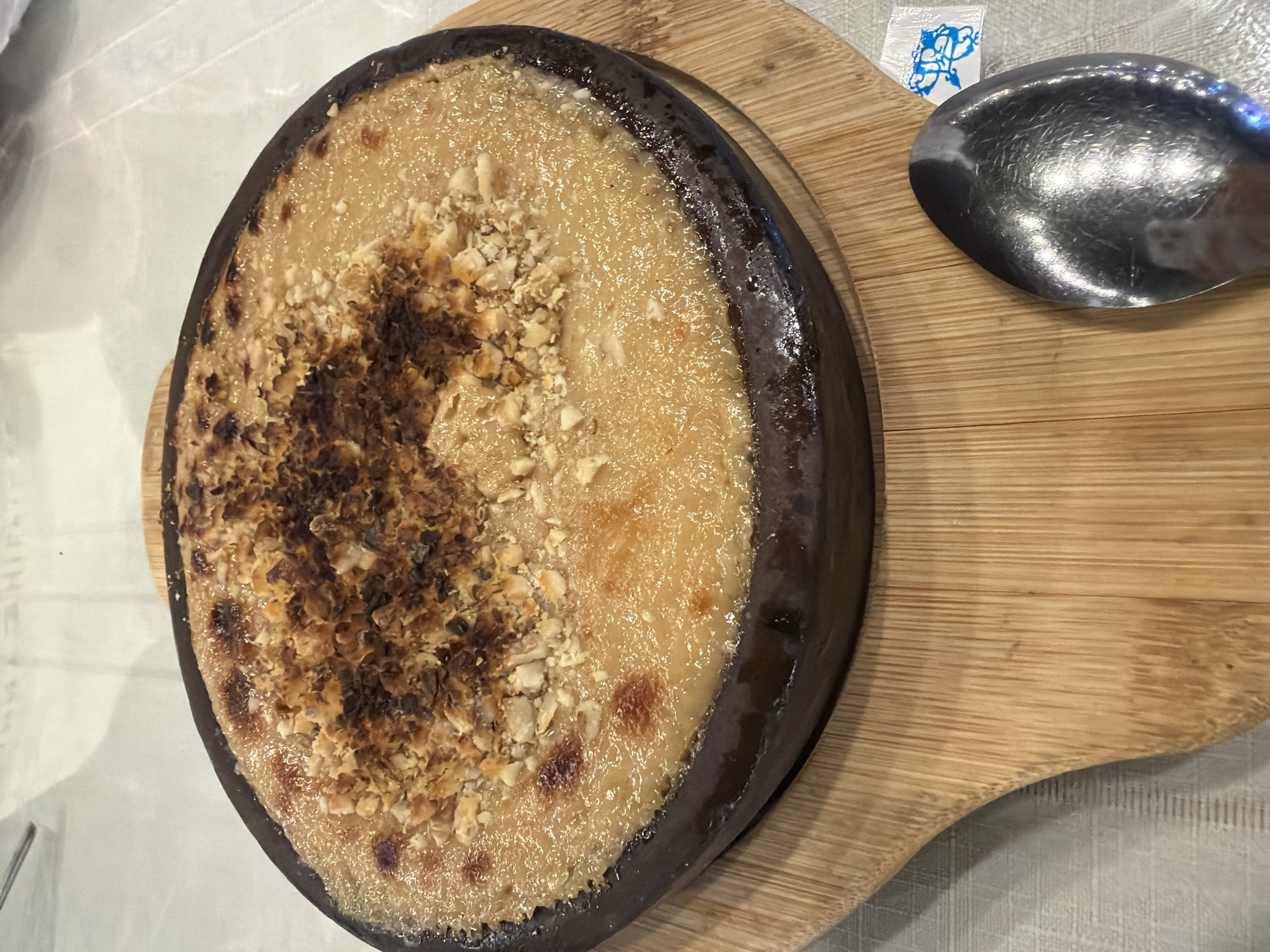 A circular, dark-brown ceramic dish containing a baked dessert is presented on a light-colored, grainy wooden board. The dessert appears to be a custard or rice pudding base with a golden-brown, slightly caramelized top, generously sprinkled with chopped nuts, some of which are charred. The dish is set on a white tablecloth. To the right of the dish, a silver spoon rests on the wooden board, and a small, white paper packet with a blue design is also visible. The image is taken from a top-down perspective, with the dessert dish occupying most of the frame. The lighting is bright, highlighting the textures and colors of the food. The scene suggests a culinary setting, possibly a restaurant in Kocaeli, Turkey, given the presence of a local delicacy.
