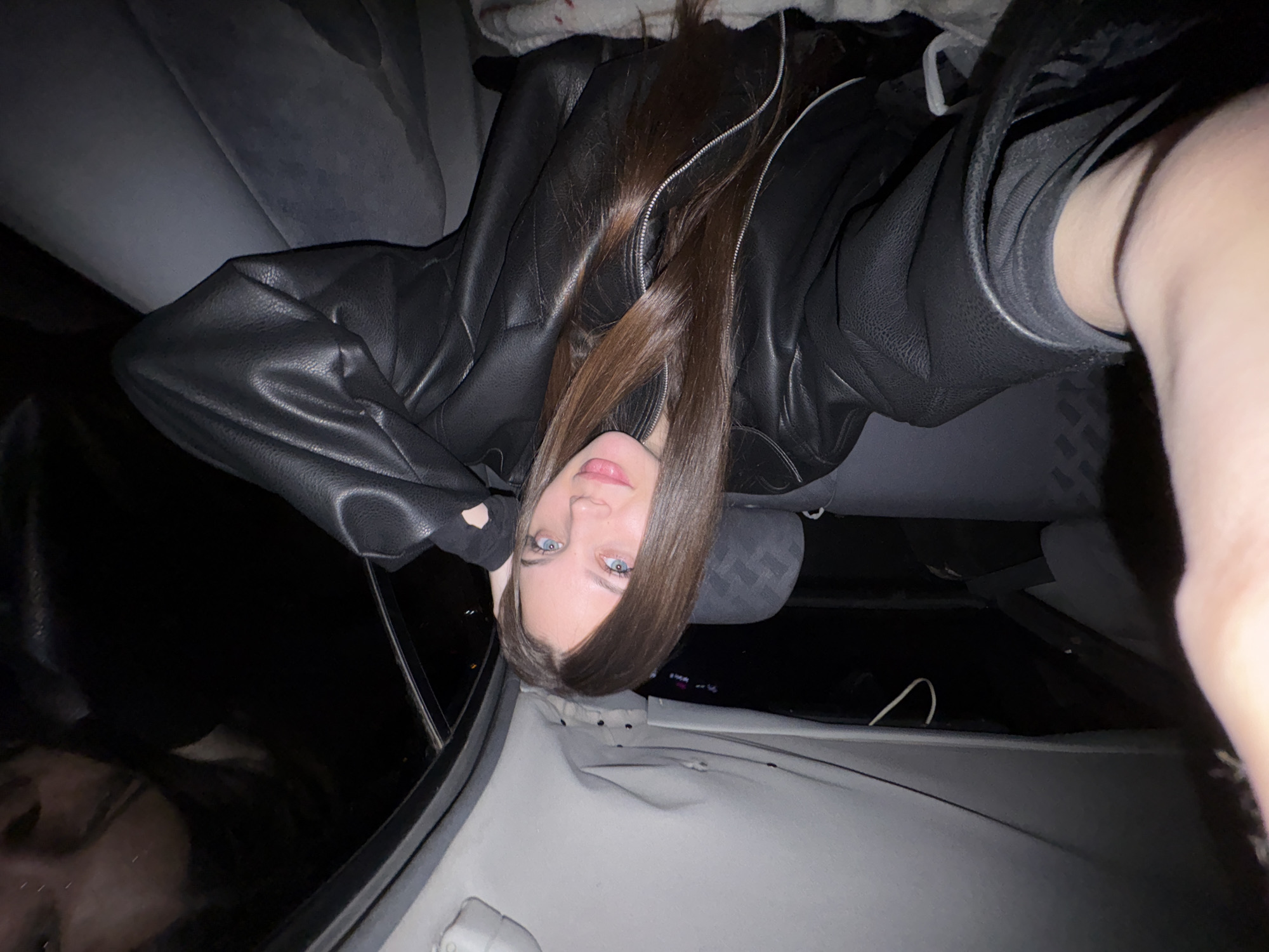 A young woman with long, straight brown hair and blue eyes is reclining in the passenger seat of a car. She is wearing a black leather jacket. The interior of the car is mostly grey, with the seat and headrest visible. Outside the car window, it appears to be dark, possibly at night. The location is not possible to infer from the image.
