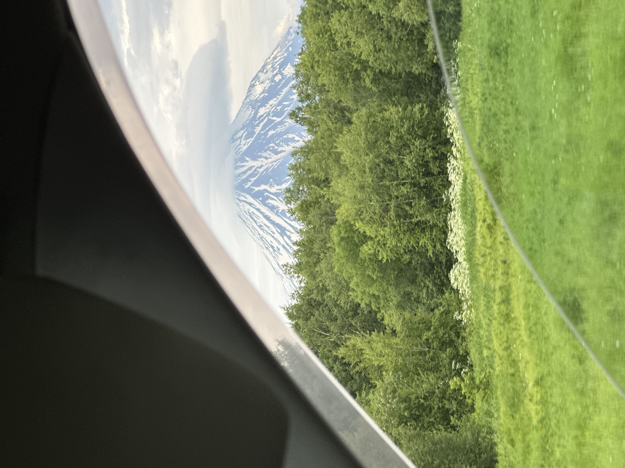 The image shows a view from inside a vehicle, looking out at a snow-capped mountain and a forest. The foreground is dominated by the dark interior of the car, with a section of the windshield and side window visible. A bright, diagonal line suggests the edge of a window frame or a reflection. Beyond this, a lush green field stretches out, dotted with small white wildflowers in the middle ground. Behind the field, a dense forest of green trees rises up. Peeking above the treeline is a majestic, snow-covered mountain with a cloud swirling around its peak. The sky is overcast with a mix of grey and white clouds. The scene depicts a natural landscape, likely outside of a populated area, and does not contain any people or specific urban elements that would help pinpoint a location in KETKINO, Russia.