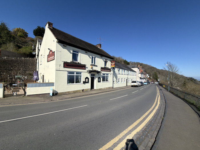 The image depicts The Fleece Inn and Malvern Hills Hotel, located on The Wyche in Malvern, Worcestershire, United Kingdom, under a clear blue sky. The primary subjects are a contiguous cream-painted, two-story building complex on the left side of a curving asphalt road. This structure features a dark tiled roof, a brick chimney, multiple white-framed windows, and a dark main entrance door. Signage prominently displays "The Fleece Inn" on a projecting board and "Malvern Hills Hotel" beneath ground-floor windows.
To the left of the main building, an outdoor patio area with tables, chairs, and a folded purple parasol is visible, enclosed by a low cream-colored wall and a black metal gate. Behind this area and the main building, a steep, vegetated hillside rises, incorporating a stone retaining wall. The asphalt road shows a single white lane marking and distinct double yellow lines along the right curb. A paved sidewalk runs along the right side of the road, separated from a descending, tree-covered hillside by a black metal fence. A white street lamp post is positioned further down the road on the right. Several parked vehicles are visible along the left side of the road adjacent to the buildings. The majority of deciduous trees on the hillsides are bare, indicating a late winter or early spring period. No individuals are present within the frame.
