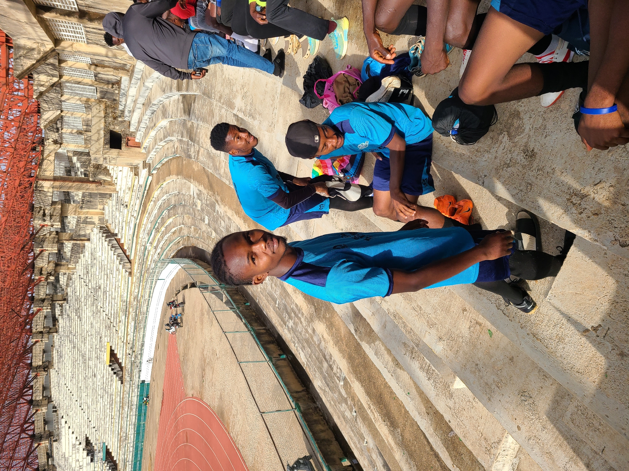 The image shows a group of young men in blue athletic wear sitting on the steps of a stadium in an unknown city. Some of the men are wearing athletic shoes. The stadium has a track and a large grandstand. The sky is clear. The lighting suggests it is daytime.