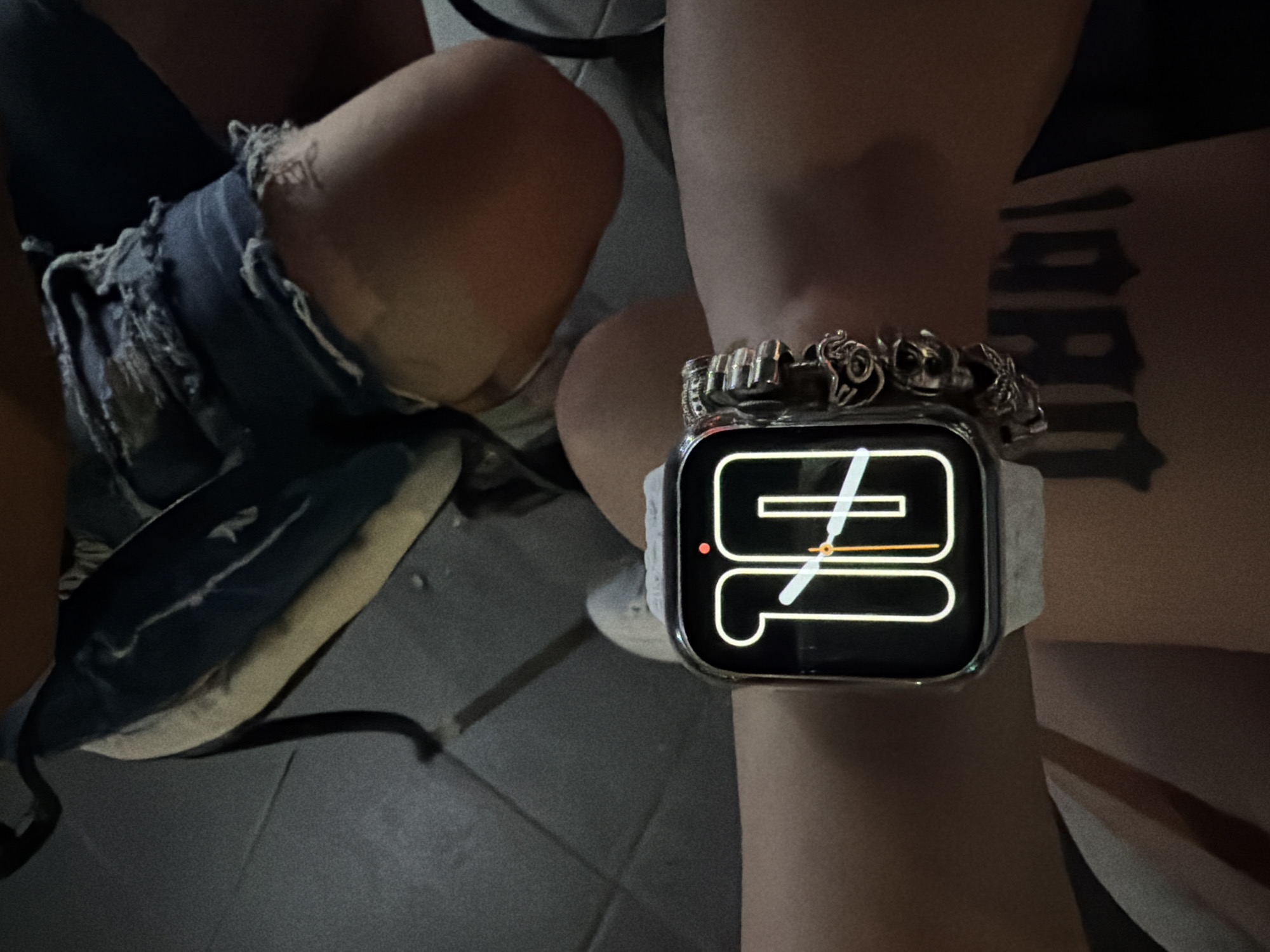 The image shows a close-up of a person's legs and arm, with a smartwatch prominently displayed on the wrist. The person is wearing ripped denim shorts. The smartwatch has a modern interface displaying a stylized clock face with numbers and hands. The watch band is white and appears to be made of fabric. The background is dark and out of focus, with a hint of a tiled floor. There's also a dark object with a light stripe that might be a bag or backpack.
A tattoo is visible on the person's thigh, featuring a stylized design that appears to be text or a graphic symbol. The lighting suggests it might be evening or indoors, and the overall mood is casual. No specific activity or event is clearly depicted beyond the presentation of the person's attire and accessories.
The location context provided is Plovdiv, Bulgaria. However, there are no visual cues in the image itself that directly indicate this specific location.