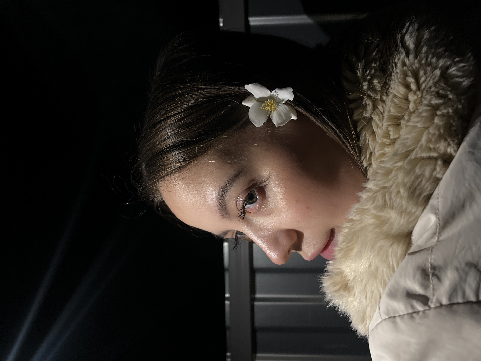 The image presents a close-up portrait of a young woman, taken at night or in a very dimly lit environment. Her head is tilted, and she gazes downwards and to her right with a gentle, introspective expression and a faint smile on her lips. She has dark hair, fair skin, and prominent eyelashes. A small, delicate white flower with visible yellow stamens is tucked into her hair above her right ear.
She is wearing a light-colored, possibly beige or off-white, quilted winter jacket with a prominent, fluffy cream-colored faux fur collar that frames her face and neck. The background is predominantly dark. To the lower left of the frame, subtle, diffused rays of light ascend diagonally, suggesting distant artificial light sources in an outdoor setting. Directly behind her, to the left of her face and below her chin, are dark, vertical panel-like structures, possibly part of a building facade or shutters. The focus is sharp on her face and the flower, highlighting the details despite the low light conditions. No text is visible in the image.