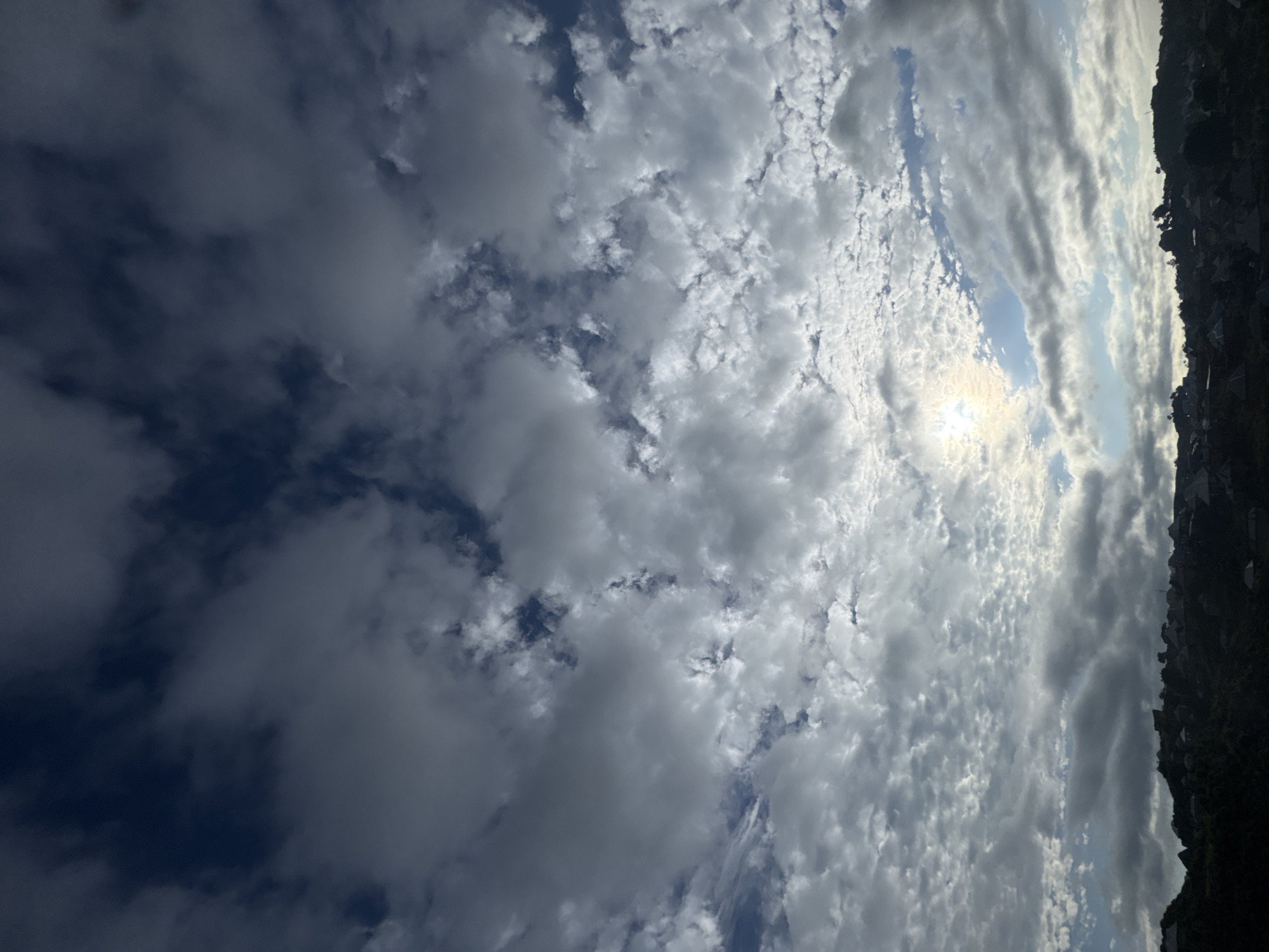 This is a photograph of a cloudy sky over a hilly landscape with a settlement on the hills. The sky is filled with various types of clouds, ranging from dark, ominous ones to lighter, scattered formations. The sun is partially visible through the clouds, casting a soft glow. The landscape below is covered in dense vegetation, and small buildings with distinct roof shapes are visible on the slopes of the hills. The overall impression is one of a natural landscape with human habitation.