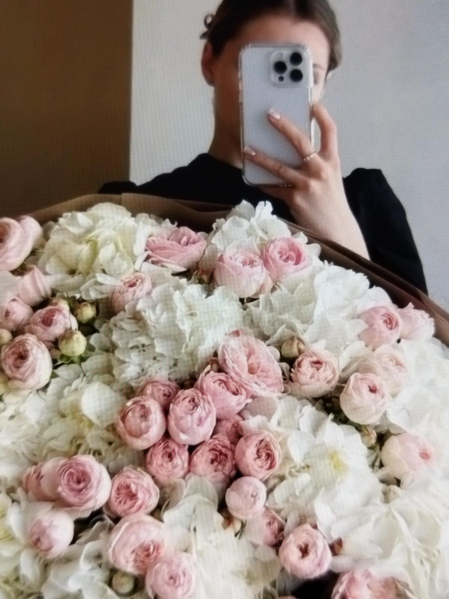 A person, partially obscured by a large bouquet of flowers, holds a smartphone in their right hand, taking a selfie. The person is wearing a black top. The bouquet consists of numerous pink roses and white hydrangeas. The background is blurred, showing a neutral wall and a wooden surface. The scene appears to be indoors. The provided metadata states the location is Beșalma, Moldova.