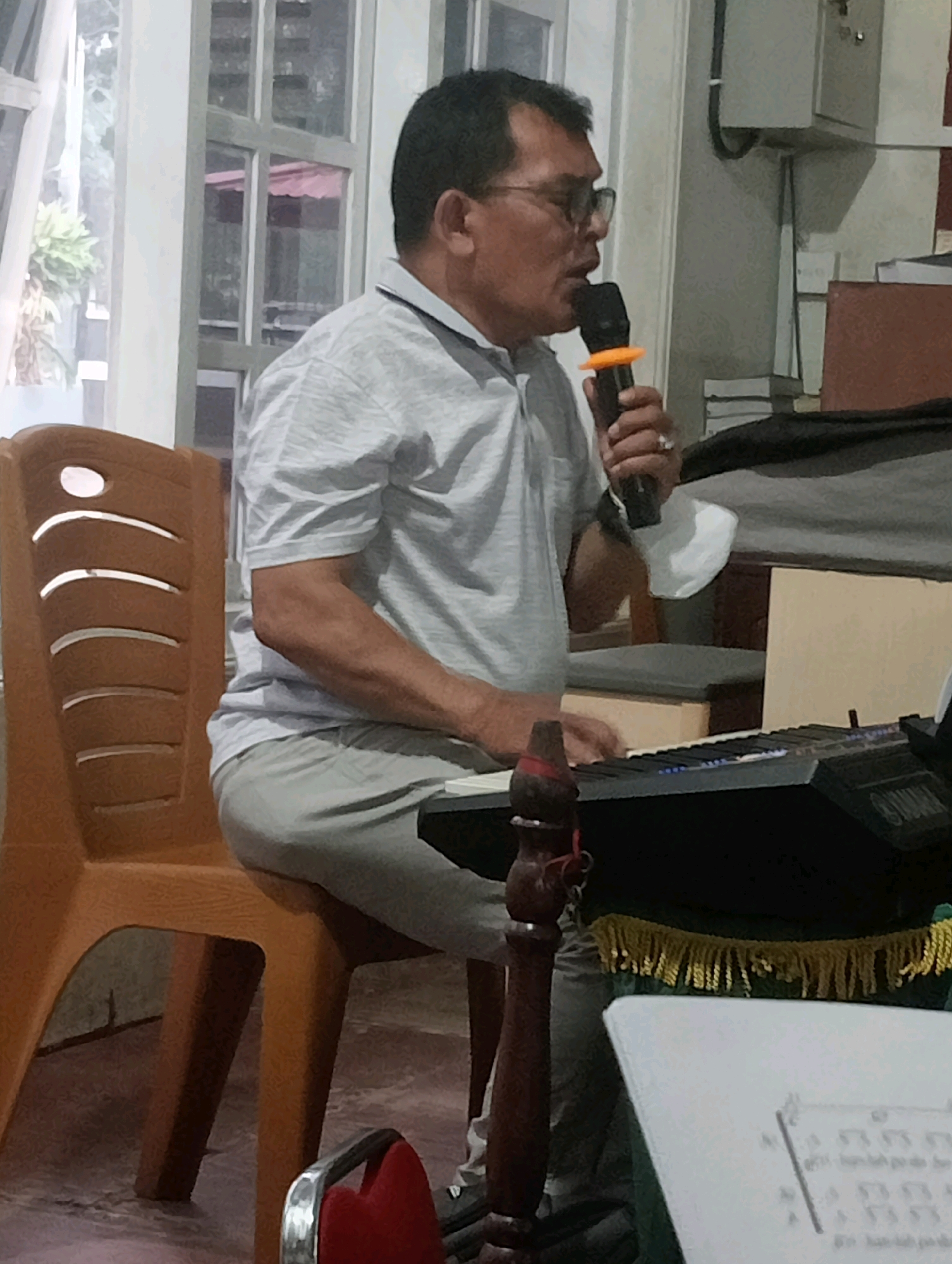 An Indonesian man wearing glasses and a light grey polo shirt is seated on a brown plastic chair and playing a black keyboard. He holds a microphone with an orange ring in his right hand, suggesting he is singing or speaking. The keyboard has "YAMAHA" branding visible. In front of the keyboard, a piece of sheet music is visible. The background includes windows with white curtains, suggesting an indoor setting. The man appears to be indoors, possibly in a community hall or church, given the arrangement of chairs and a keyboard. The image does not provide enough information to determine the specific location within Jambi, Indonesia, beyond it being an indoor setting.