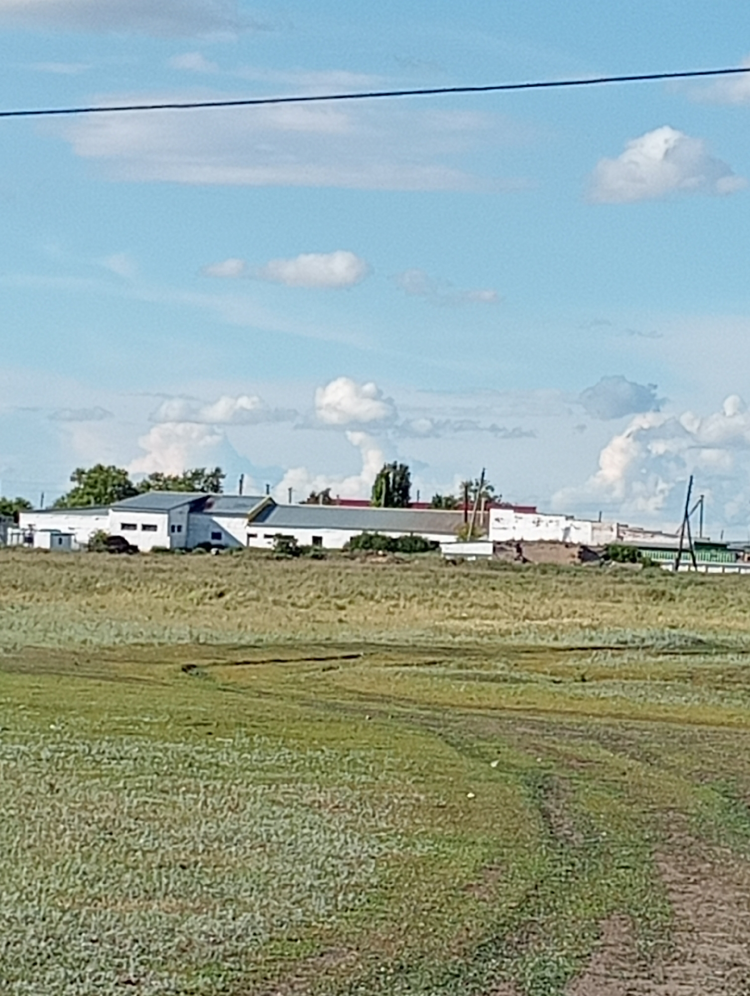 The image shows a wide view of a rural landscape under a blue sky with scattered clouds. In the middle ground, there is a collection of white, low-rise industrial or farm buildings with dark roofs. Some of these buildings appear to be interconnected. Trees and vegetation are visible around and behind the buildings. The foreground consists of a grassy field with some tire tracks or pathways visible. A dark line, possibly a power line, runs horizontally across the top of the image. The overall scene depicts a somewhat desolate, possibly agricultural or industrial area in a rural setting.