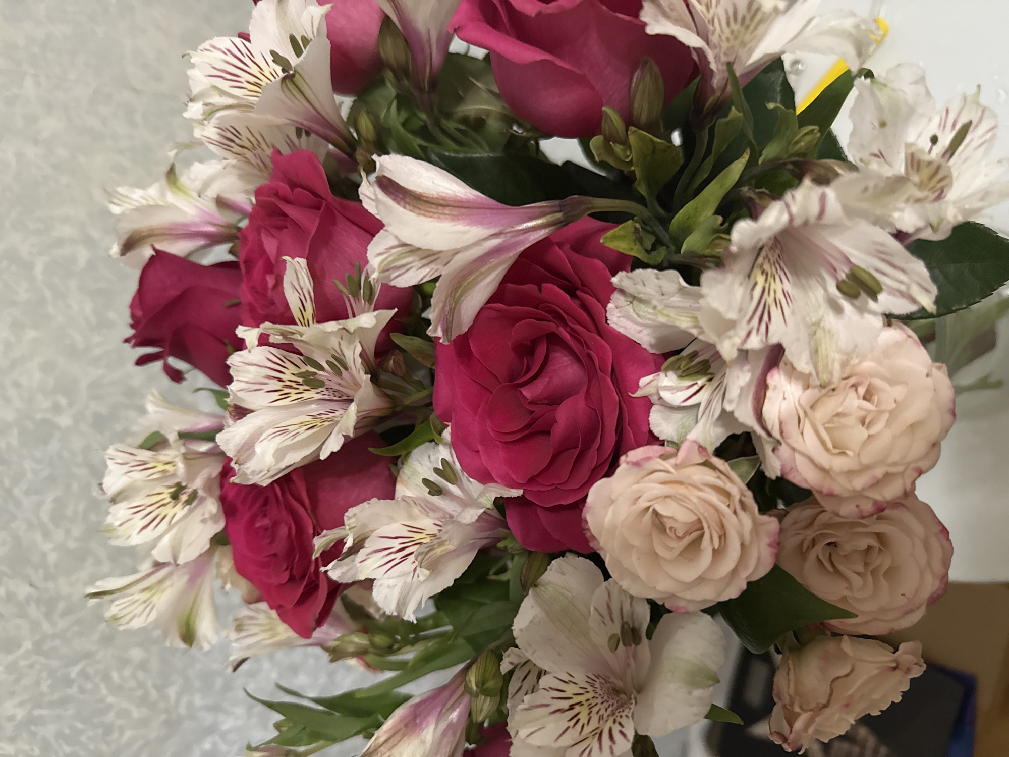 The image shows a bouquet of flowers with pink roses and white Alstroemeria. The roses are primarily in shades of pink, with some appearing a deeper, almost reddish-pink, and others a lighter, blush pink. The Alstroemeria flowers are white with delicate purple or brown striations. Green foliage is visible between the blooms. The background appears to be a light, textured surface, possibly a wall. There is no indication of a specific location, especially in Saint Petersburg, Russia, nor are there people or actions visible in the image. The only objects are the flowers and what appears to be a wall.