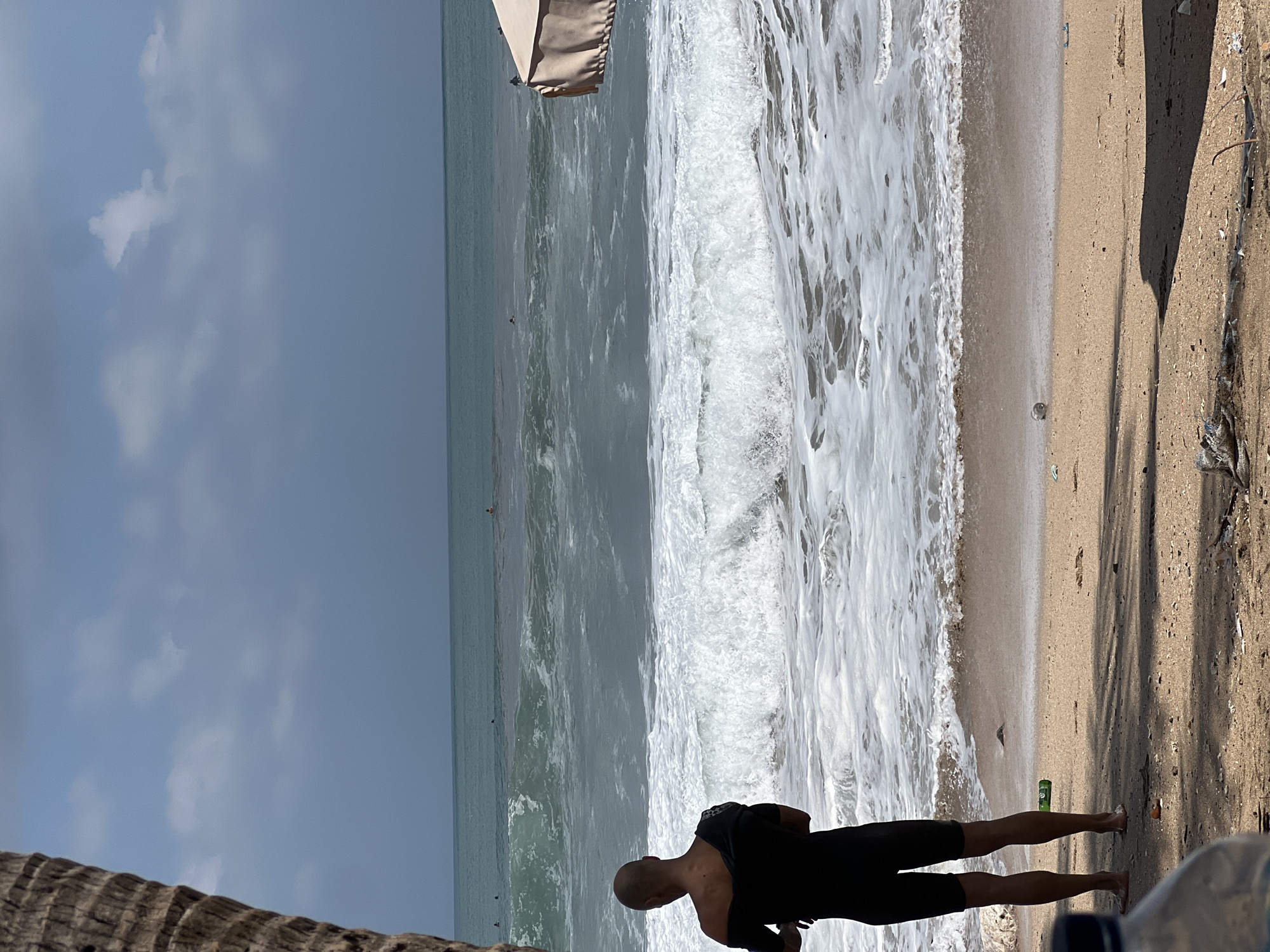 A person, viewed from behind and from the waist down, stands on a beach in Kuta, Indonesia, by the ocean. The individual is wearing a black wetsuit, which they are in the process of removing. The person's legs and lower torso are visible, and their arms are holding the sides of the wetsuit, pulling it down. The beach is composed of sand, with some debris scattered across its surface, including plastic bottles and what appears to be trash. The ocean is active with waves breaking on the shore, creating a foamy white surf. Several other individuals can be seen in the water, some appearing to be surfing or paddling. In the background, the sky is overcast with patches of blue and clouds. The lighting suggests it is daytime, likely in the afternoon. A beach umbrella is partially visible on the right side of the frame. A portion of a palm tree trunk is visible on the far left side of the image.