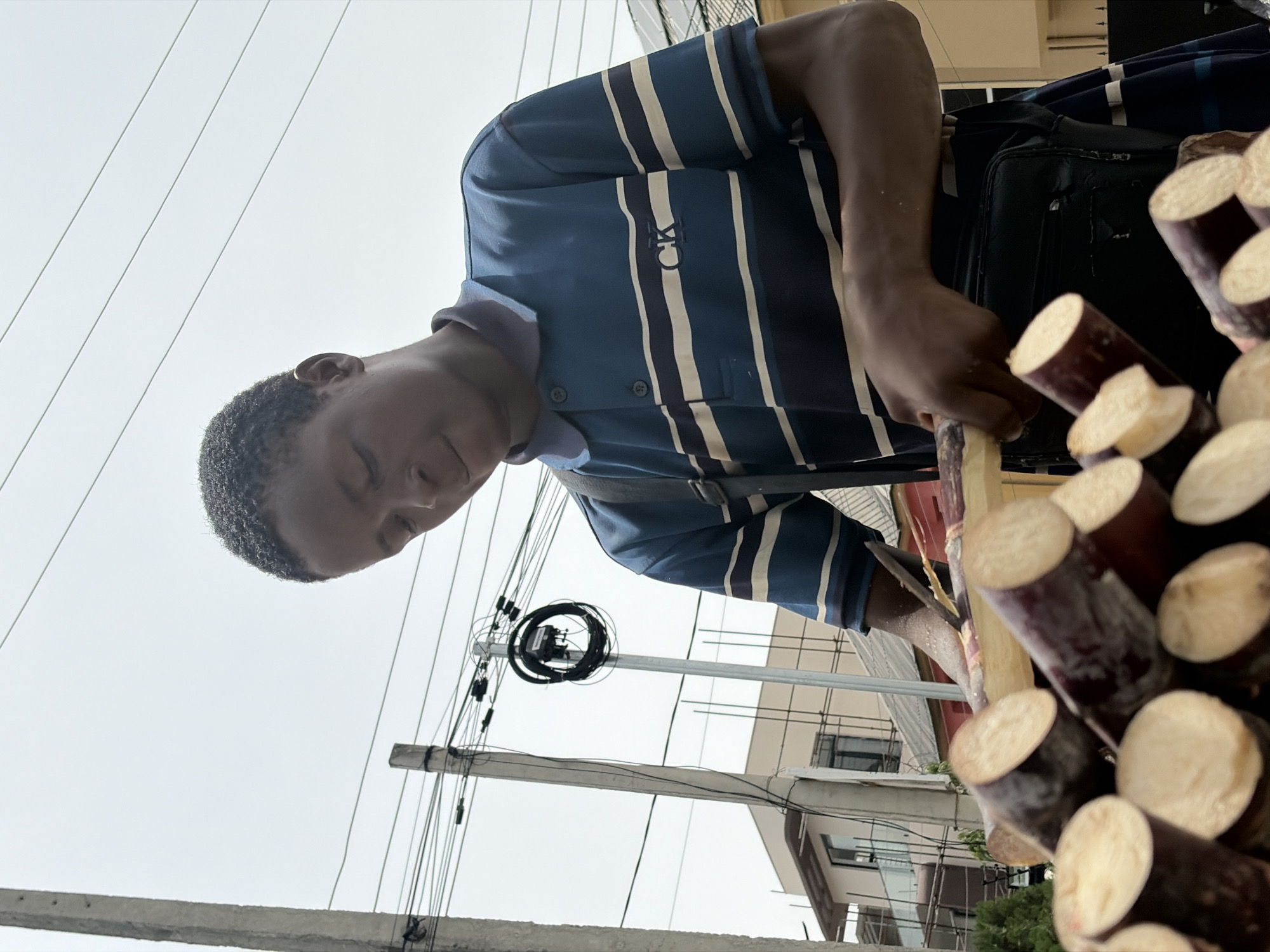 The image depicts a man in Igboefon, Nigeria, preparing sugar cane. He is wearing a blue and white striped polo shirt with a visible "CK" logo. He is using a knife to peel a stalk of sugar cane. A large pile of sugar cane stalks, cut and peeled, is prominently displayed in the foreground. The background includes overhead power lines, concrete poles, and buildings typical of an urban environment in Igboefon. The sky is overcast.