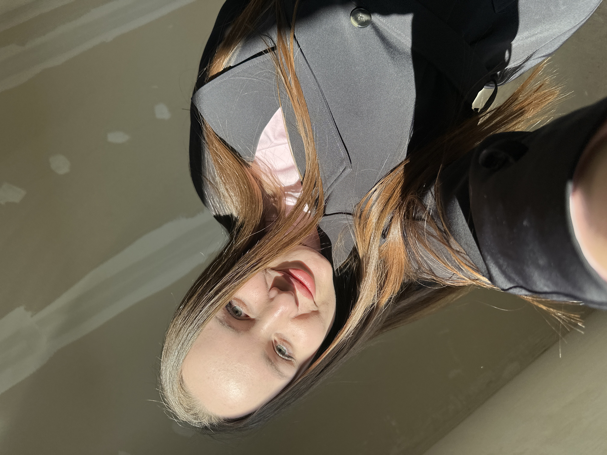 A low-angle selfie captures a young female indoors, presumed to be in Pervomaysky, Russia. The subject, positioned centrally and tilted slightly, has long, straight, brown hair with lighter highlights and a fair complexion. She displays a slight smile, revealing light-colored (likely green or blue) eyes and pink-tinted lips. She is wearing a dark grey or black blazer with a prominent lapel and a visible grey button, over a light pink garment. Strong, direct sunlight illuminates her face and hair from the left, casting distinct shadows on her right side and the background. The background consists of a plain, light beige wall showing visible patches or texture variations, suggestive of an interior space under construction or renovation. The fingers of the hand holding the camera are partially visible in the bottom-left corner of the frame.
