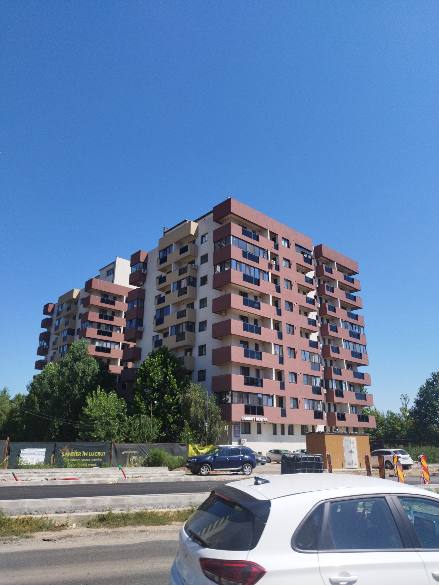 The image captures a clear, sunny day in Bragadiru, Romania, with a modern apartment building dominating the scene. The building, with its distinctive red and beige facade and numerous balconies, is a central focus. In the foreground, a construction site is visible with new roadwork in progress, indicated by asphalt and concrete barriers. Several vehicles, including a white car in the immediate foreground and a blue SUV, are present. Signs for "SANTIER IN LUCRU!" (Worksite!) and "CABINET DENTAR" (Dental Office) are discernible on or near the building, providing context for the location. Lush green trees surround the construction area and the building, contrasting with the urban infrastructure. The sky is a vibrant, cloudless blue.