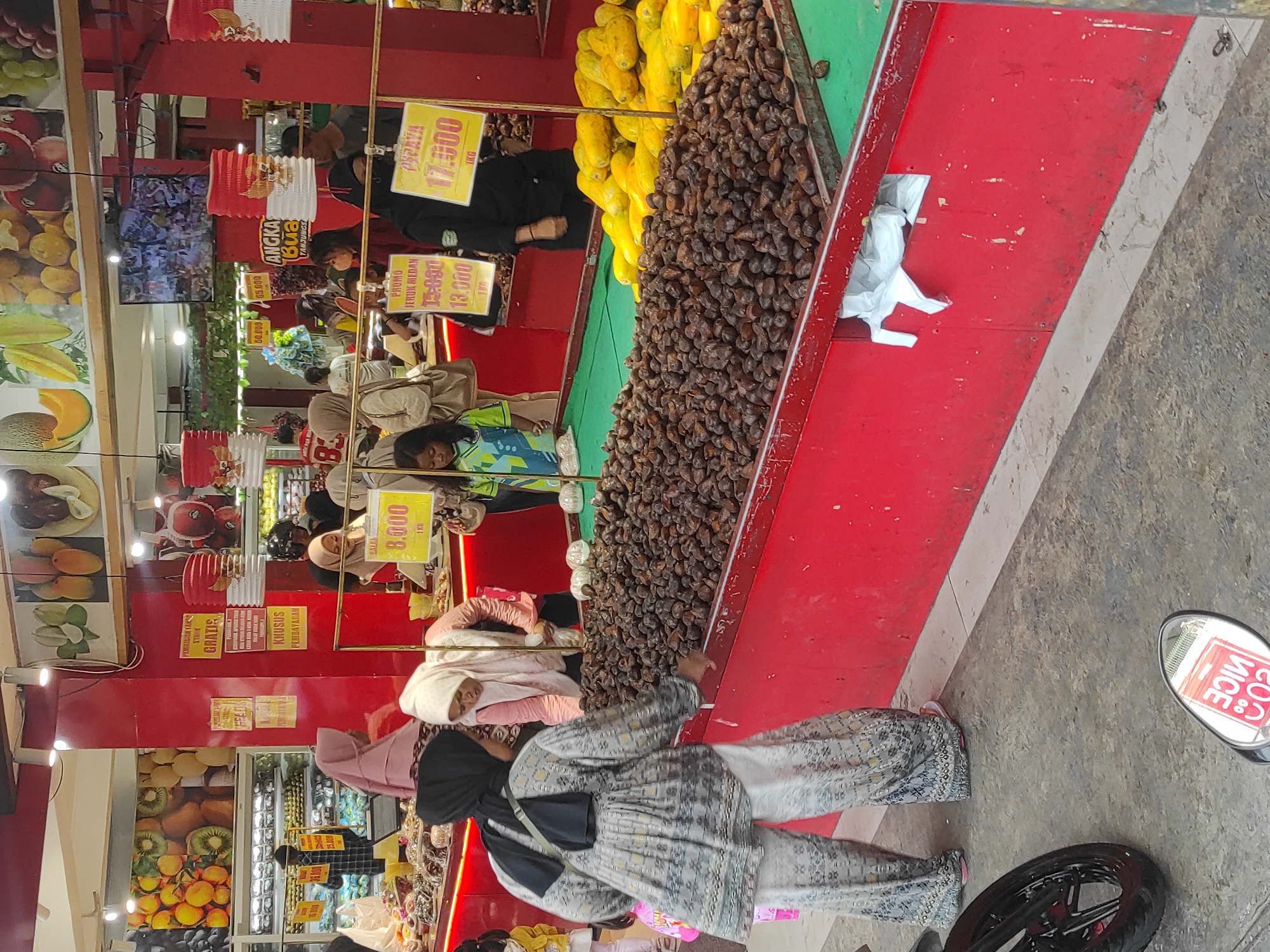 The image depicts a bustling fruit market scene, likely in Sumedang, Indonesia. In the foreground, two women are interacting near a large display of brown, nut-like fruits. One woman, wearing a black hijab and a patterned gray outfit with wide-legged pants, appears to be examining the fruit. The other woman, in a pink hijab and a pink top, stands behind the counter. In the middle ground, several other shoppers, including a young girl in a green and blue shirt, are browsing the produce. The market stall is adorned with red trim, and yellow price signs are visible. Behind the shoppers, more fruits are displayed, including what appear to be papayas. Red and white paper lanterns hang from above. The background shows more stalls and displays of fruit, suggesting a well-stocked market. The overall environment appears to be indoors or under a covered area, with overhead lighting illuminating the scene. A motorcycle wheel is visible in the lower left corner of the image.