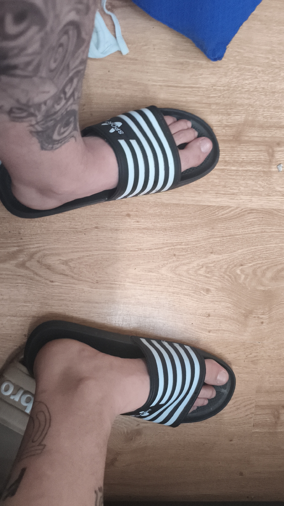 A high-angle shot reveals two feet, each adorned with a black and light blue striped slide sandal, positioned on a light brown wooden floor. The feet are fair-skinned, with visible tattoos on the lower legs. The upper leg in the frame displays a large, dark tattoo. The sandals feature a sporty design with multiple light blue stripes against a black base. The toes of each foot are visible, peeking out from beneath the sandal straps. The floor is free of clutter, with a blue cloth visible in the upper right corner. The photograph's location, Nowy Tomyśl, Poland, is included for contextual purposes only and not visually apparent in the image.