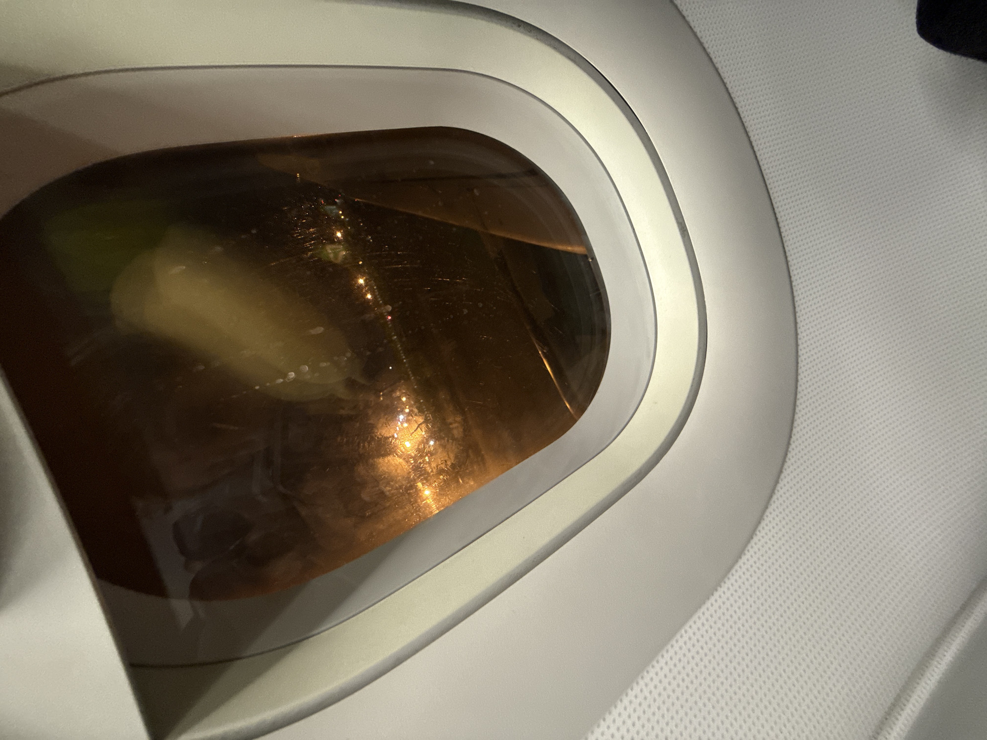 The image shows a view from an airplane window at night, overlooking the city of Ob, Russia. The window, with its characteristic oval shape and white frame, is slightly smudged, reflecting the interior of the aircraft. Through the window, the city lights of Ob are visible, forming a scattered pattern of warm yellow and white dots against the dark background. Some of the lights appear to be along roads or industrial areas, suggesting urban or suburban development. The reflection in the window also shows blurry shapes that could be parts of the aircraft's interior or possibly other passengers. The overall impression is one of a nighttime aerial view of a city, captured from the confined perspective of an airplane.