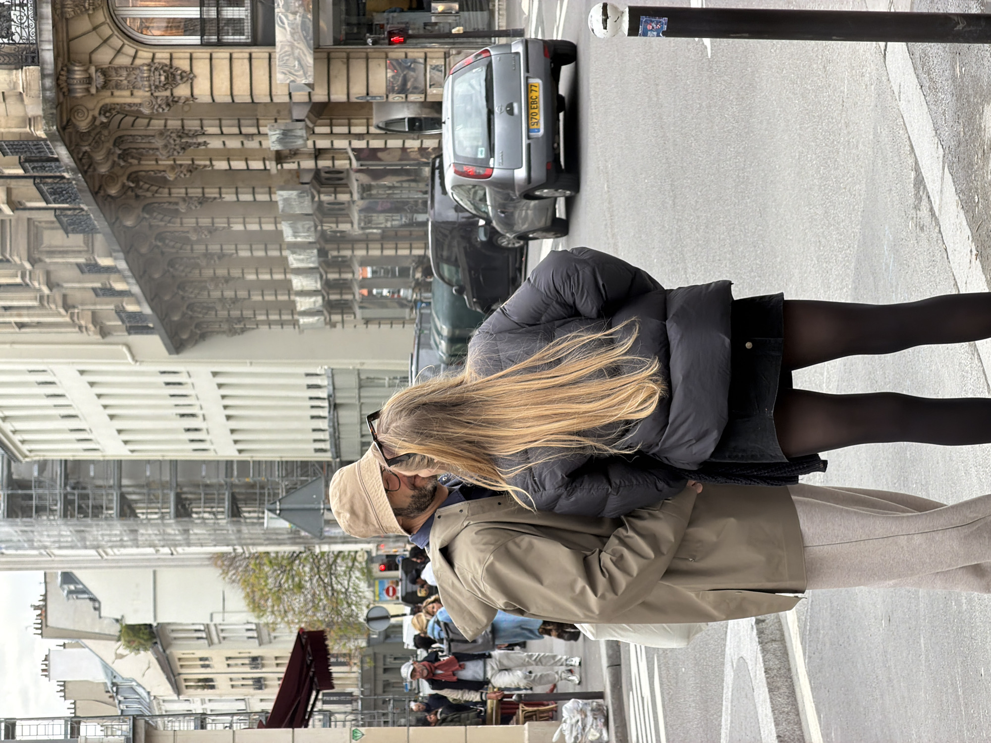 The image shows a couple kissing on a sidewalk in Paris, France. The woman has long blonde hair and is wearing a black puffer jacket and black tights. The man is wearing a tan jacket and light-colored pants. The background includes Parisian buildings, traffic, and pedestrians. A grey car is parked along the street.