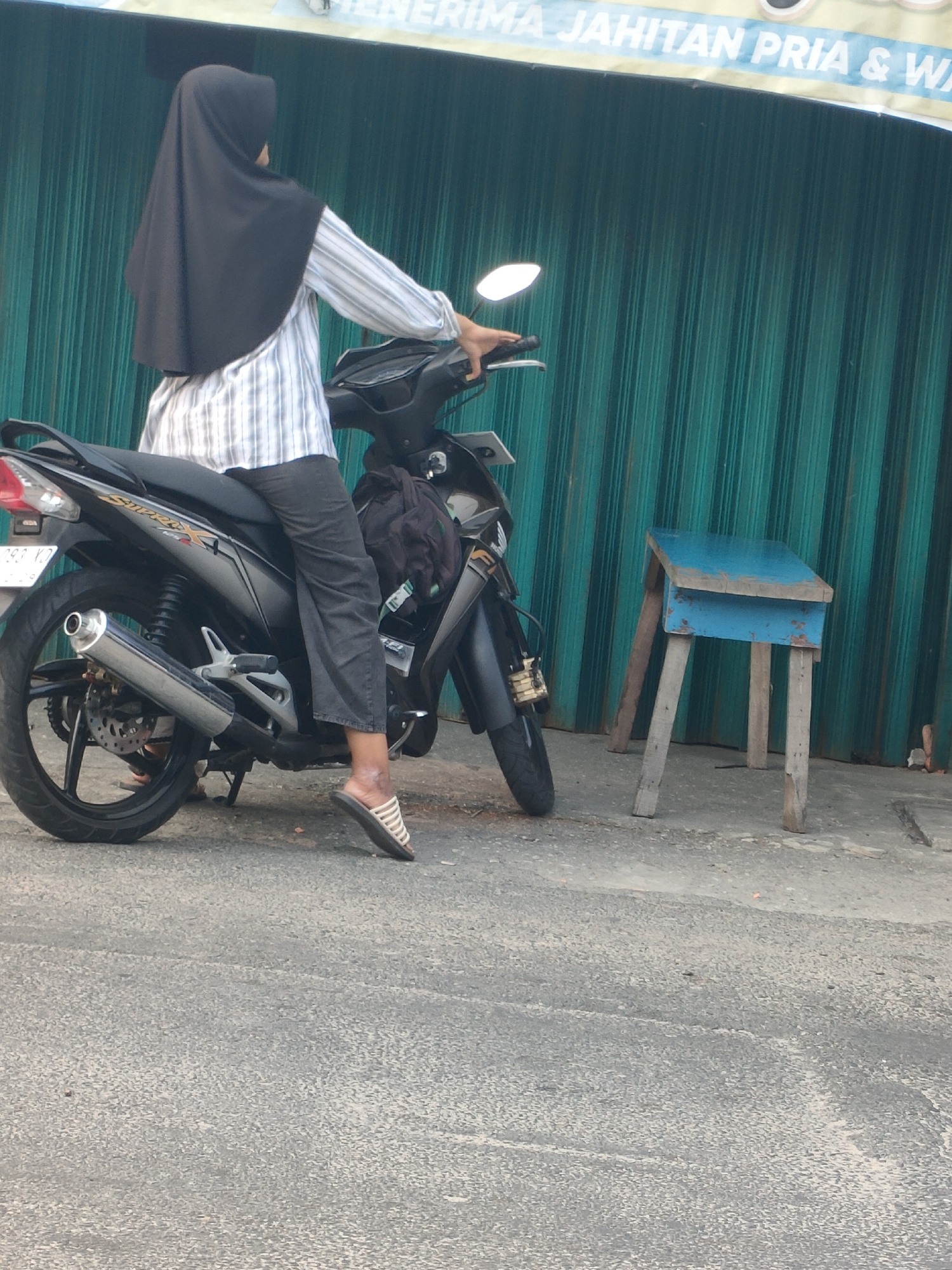 A person wearing a black hijab and a striped long-sleeved shirt is seated on a black motorcycle. The person's legs are crossed, with their right foot wearing a white sandal with horizontal stripes. The motorcycle has a license plate that appears to be from Indonesia. To the right of the motorcycle is a small, rustic blue wooden stool with four legs. Behind the motorcycle and stool is a teal-colored corrugated metal shutter door. Above the shutter door, a banner with text that is partially visible, including the words "JAHITAN PRIA & WA" is displayed. The ground is paved with asphalt. The scene appears to be in Pekanbaru, Indonesia.
