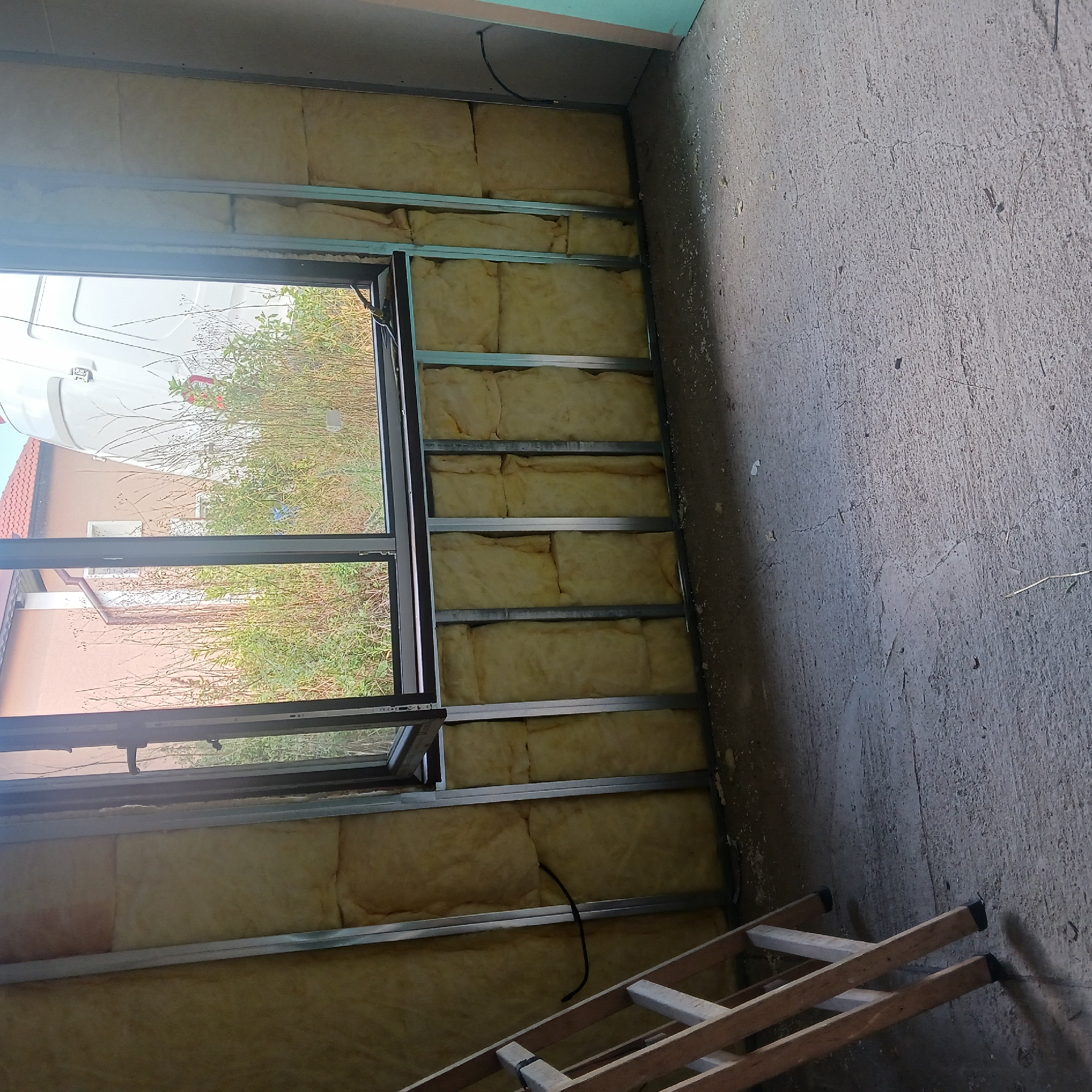 The image shows an interior room under construction, likely in the city of Ostra Mogila, Bulgaria. The walls are partially covered with yellow insulation material and metal framing, indicating a construction or renovation phase. A window provides a view of an overgrown outdoor area with some vegetation, a house with a red roof, and a white vehicle, possibly a van or a camper. A wooden ladder is placed against the left wall. The floor appears to be concrete, with some debris and dust present. A black electrical cable is visible on the insulated wall. The overall scene suggests a work in progress.